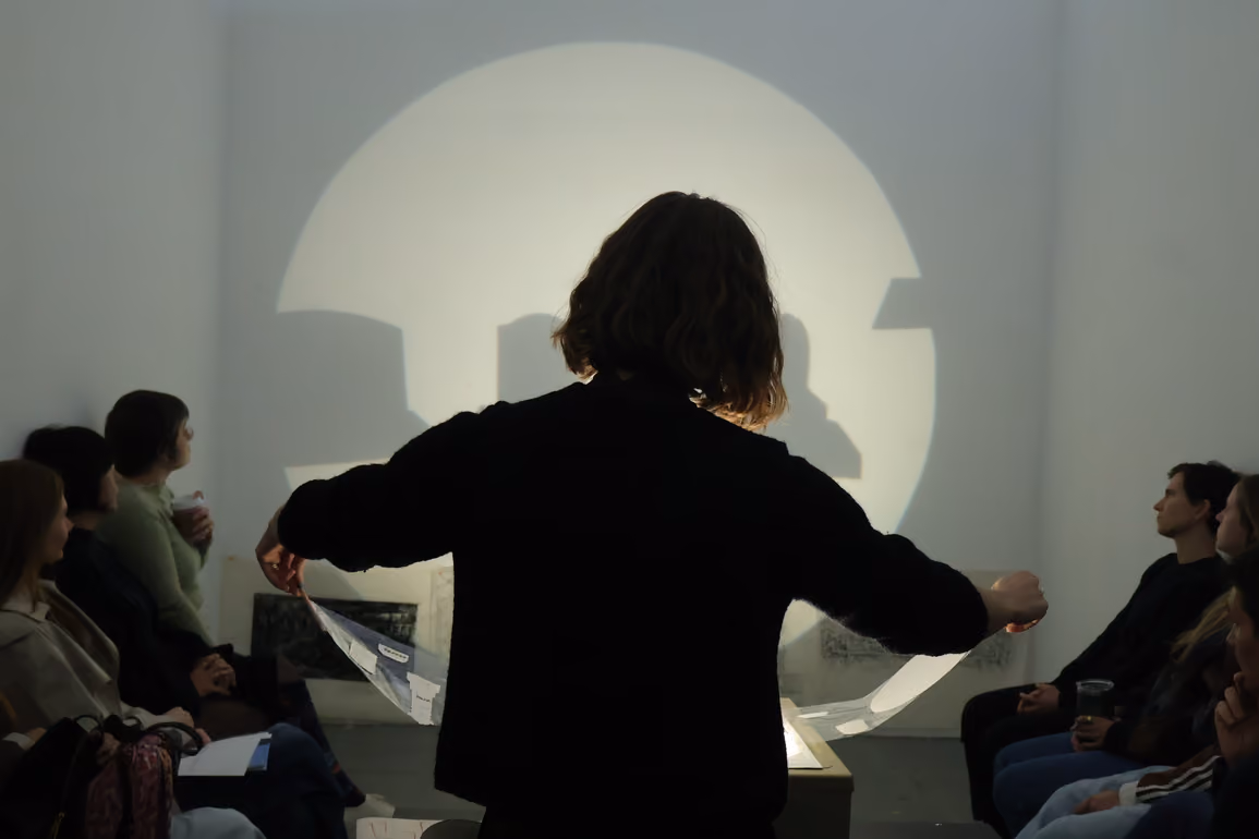 Silhouette view from behind a presenter holding a document before a circular light beam projected onto a wall, surrounded by an seated audience.