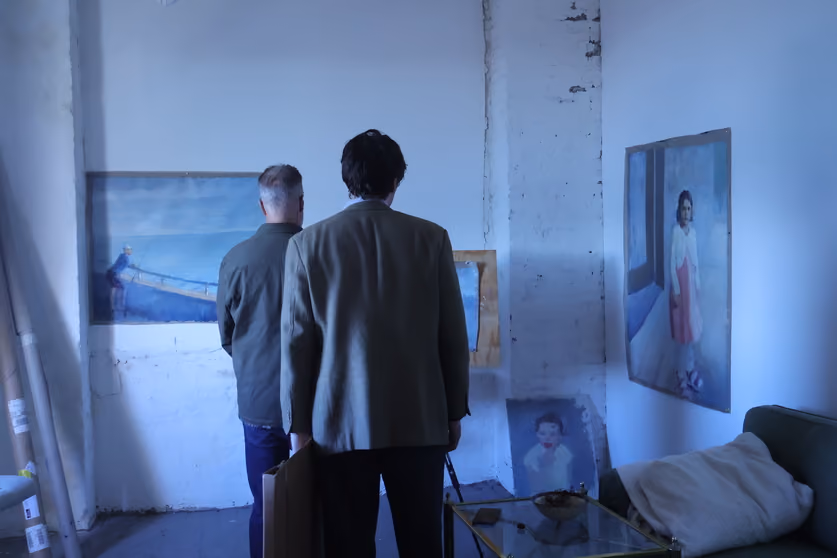 Two men viewed from behind as they observe figurative and landscape paintings mounted on the walls of a cozy studio space.