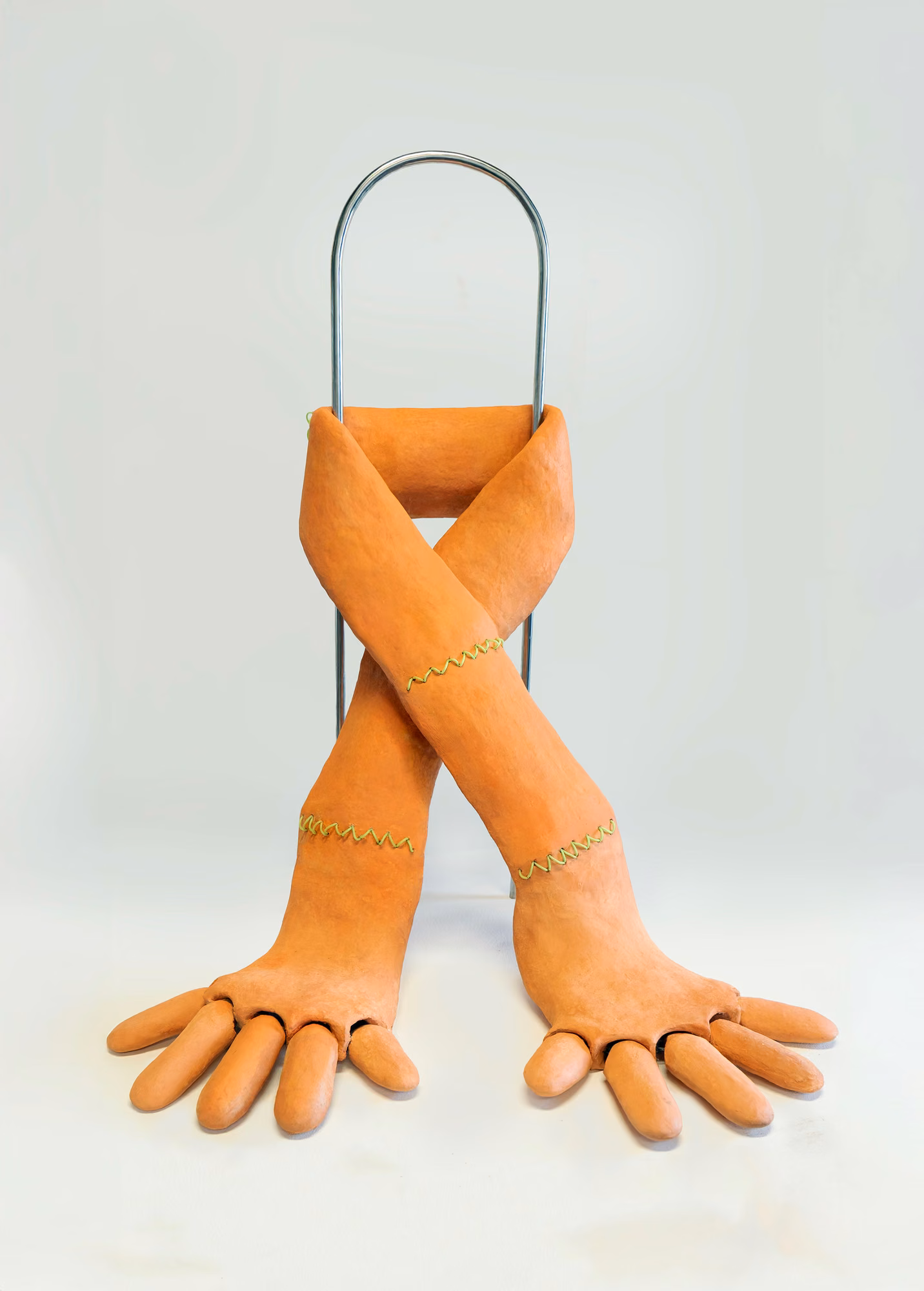 Work by Ping Wang (X): orange ceramic pair of crossed arms with finger-like hands, stitched in yellow, draped over a metal arch.