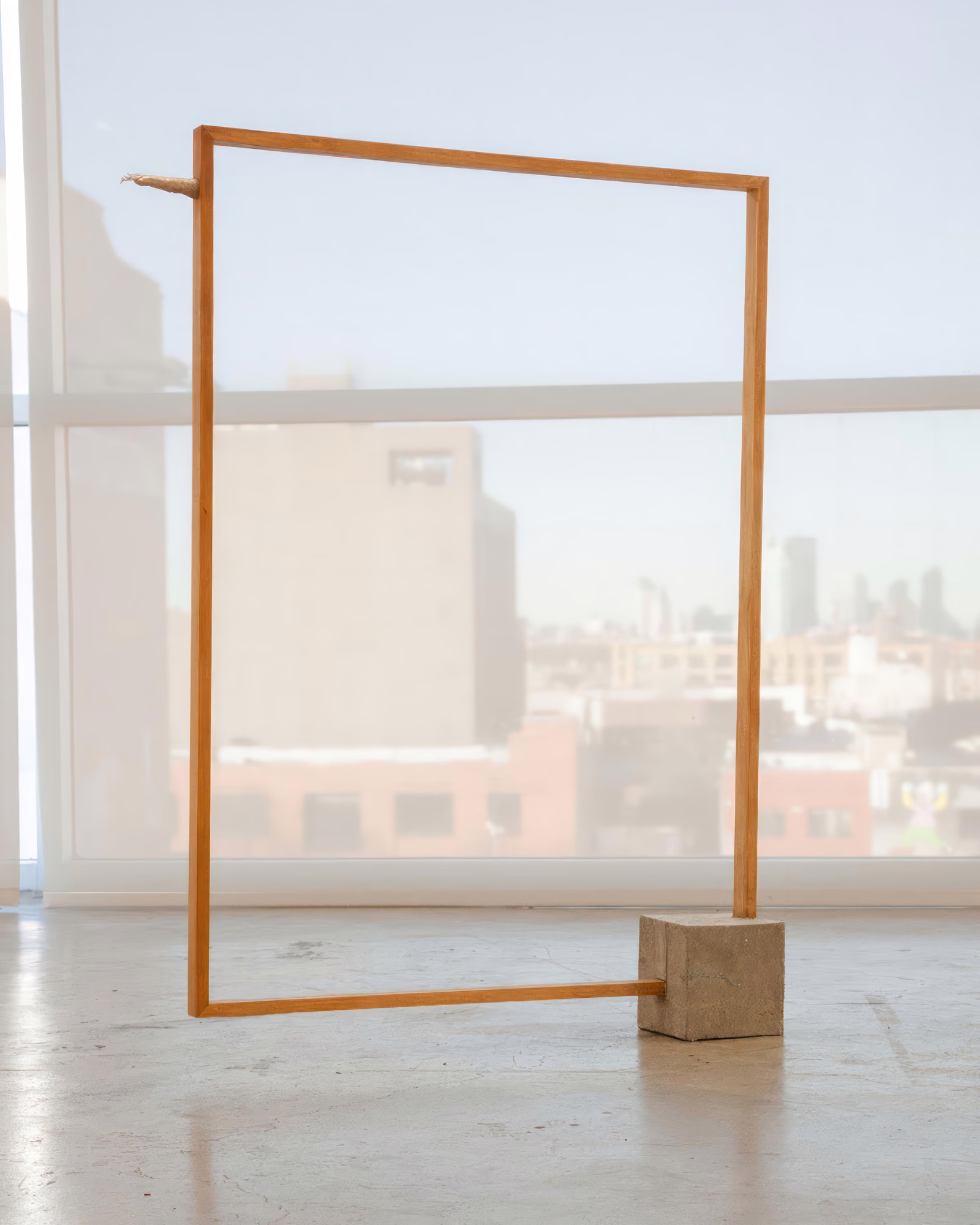 Work by Ping Wang (X): open wooden rectangular frame standing on a small block before a large window with a city view.