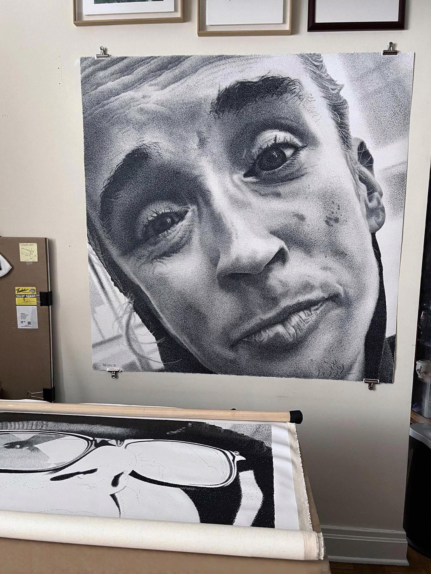 Work by Richard Masters: large black-and-white portrait drawing of a face pinned to a wall, with a rolled drawing of glasses below.