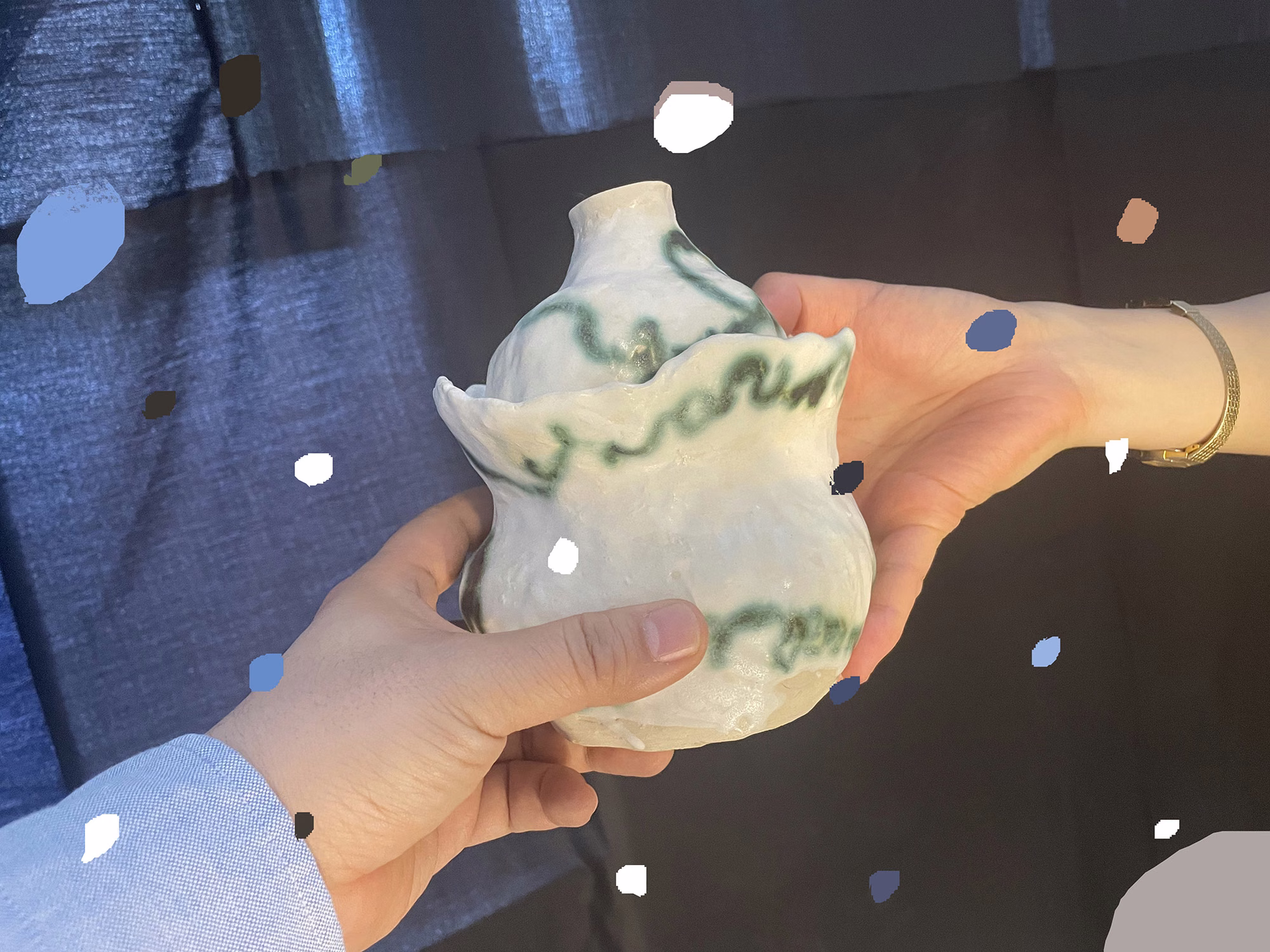 CONFLUENCE UNDER BRINE: two hands hold a small white ceramic vessel with green wavy lines, against dark blue fabric dotted with white and blue spots.
