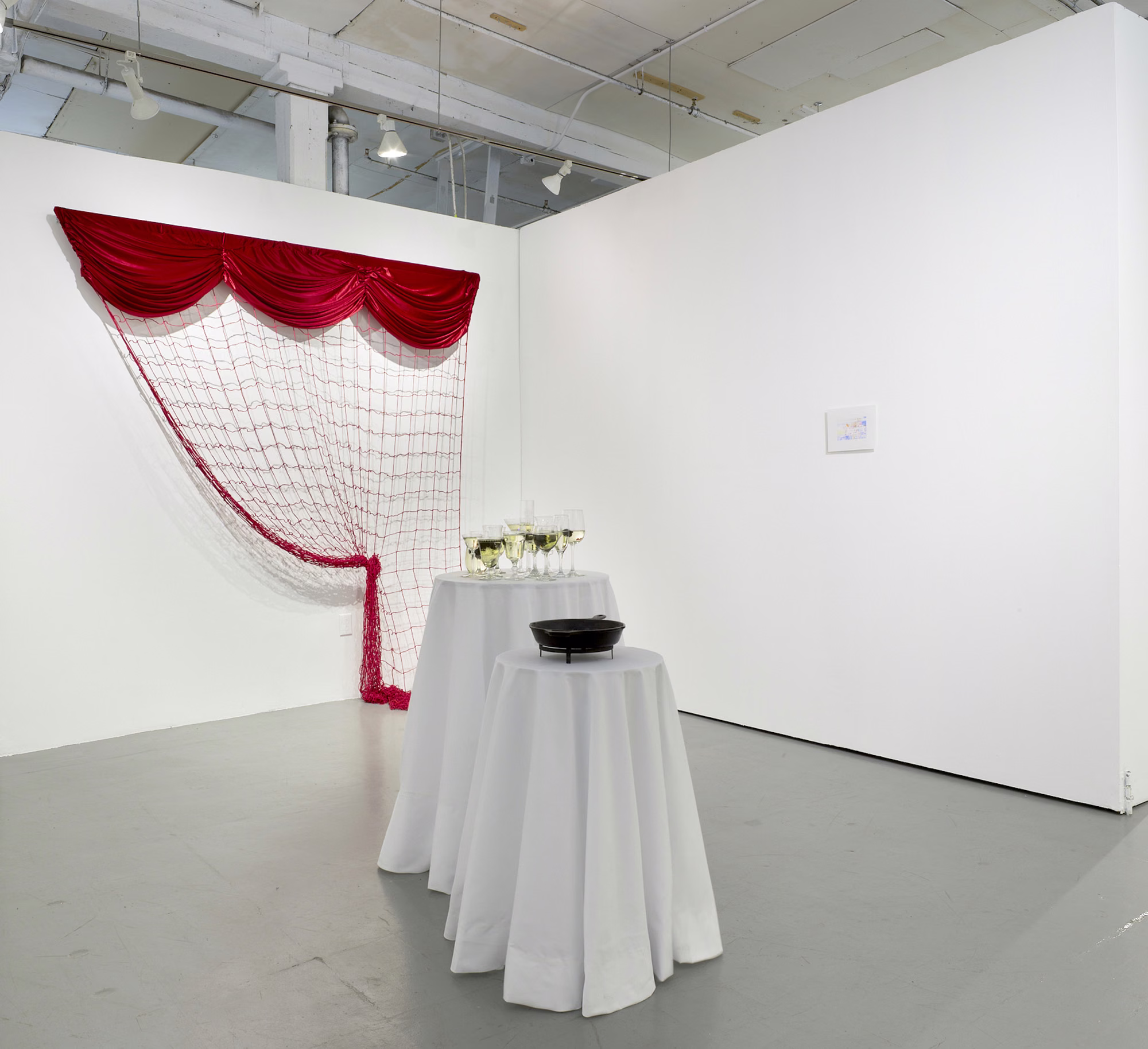 Installation view of Toward a Sentence: a red curtain valance and netting hang in a gallery corner above two tables with white cloths and flower arrangements.