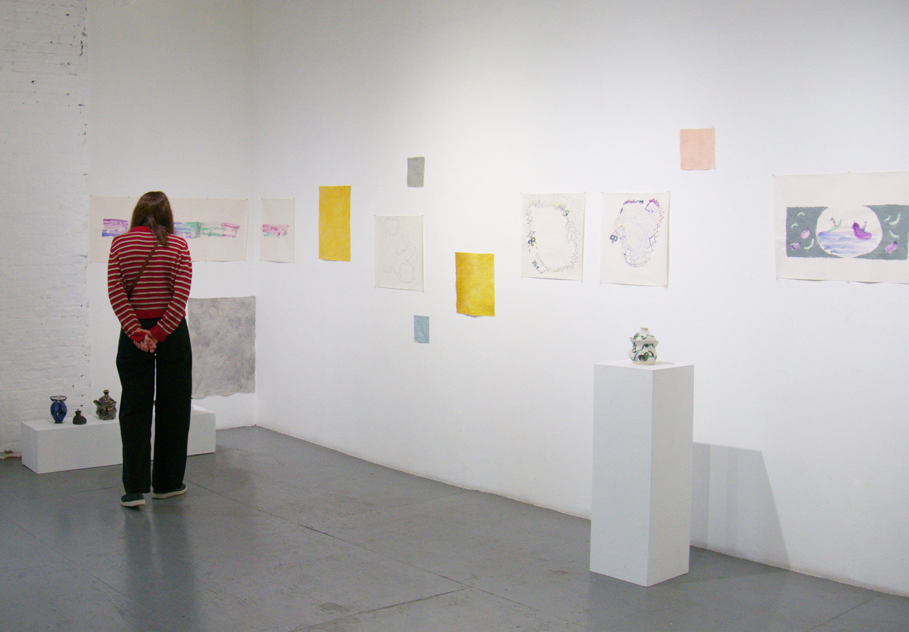Yoshie Sugito, 2026. Image description: BONE ASH, MEMORY HOLES, THE RECIPES: a visitor in a red-and-white striped sweater views drawings and ceramic works in a white gallery space.