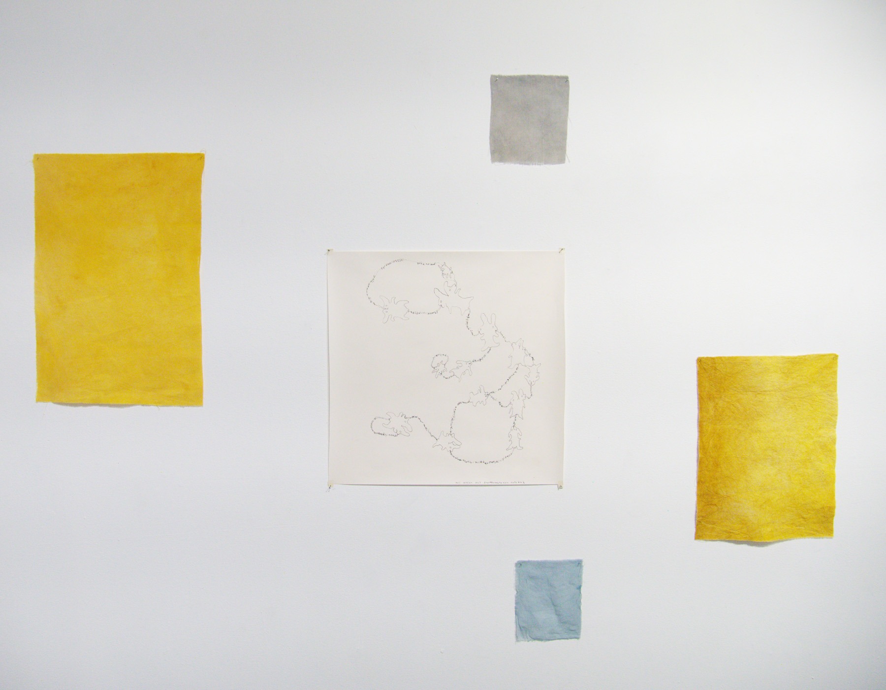 Yoshie Sugito, 2026. Image description: Installation view of BONE ASH, MEMORY HOLES, THE RECIPES: a central paper drawing surrounded by yellow, gray, and blue fabric swatches on a wall.