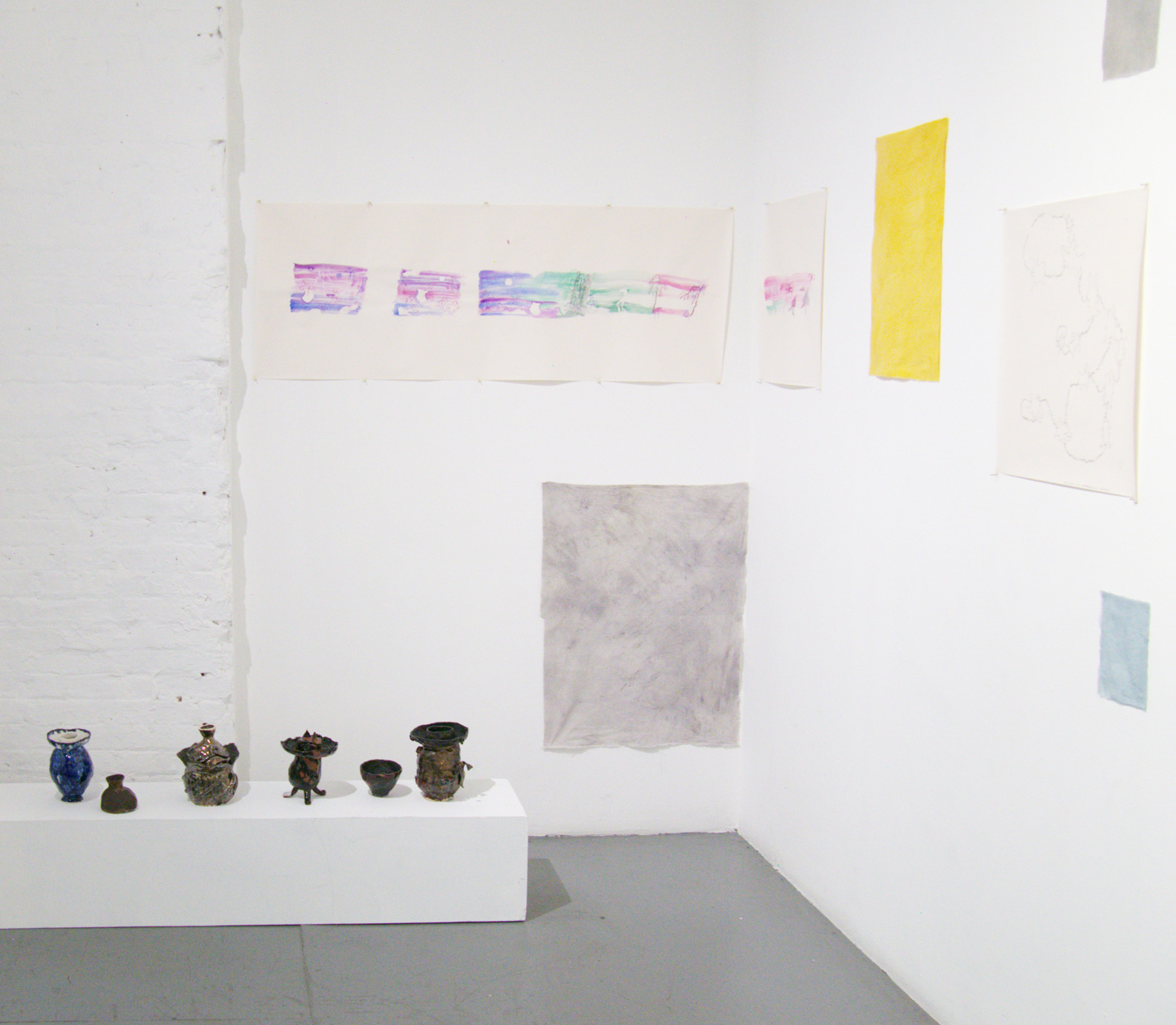 Yoshie Sugito, 2026. Image description: Installation view of BONE ASH, MEMORY HOLES, THE RECIPES: dark ceramic vessels on a low white pedestal near works on paper and fabric.