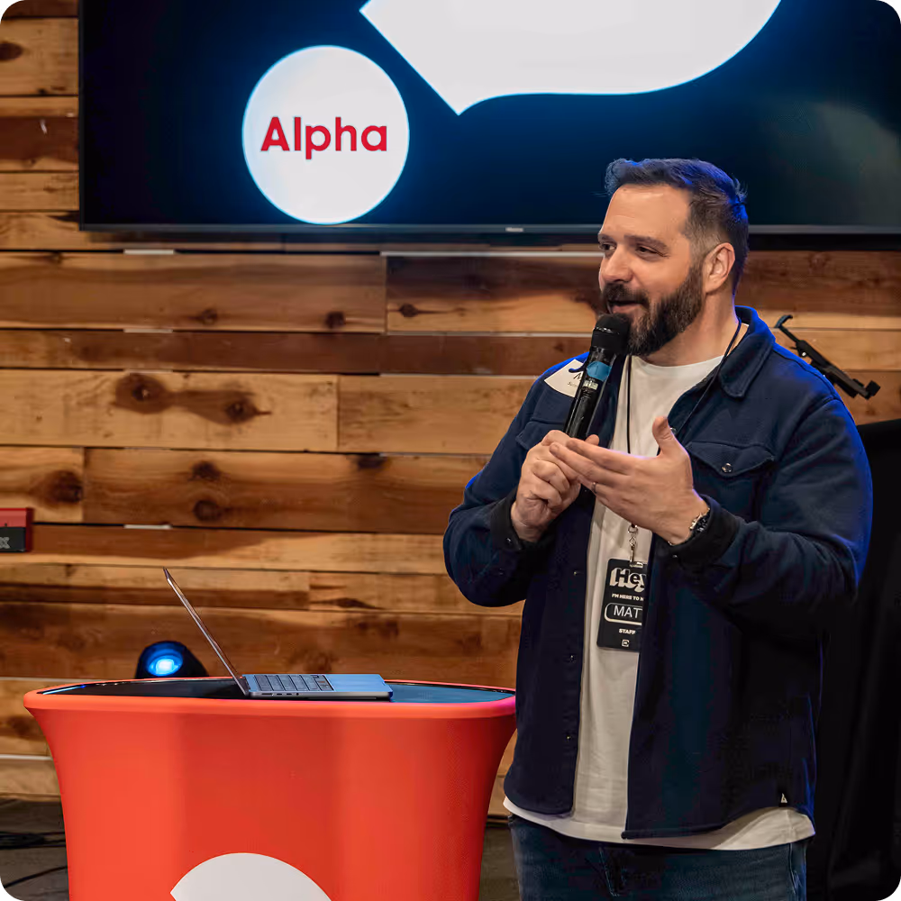 Man with beard speaking into a microphone next to a red podium with a laptop, in front of a wooden wall and a screen displaying the word 'Alpha'.