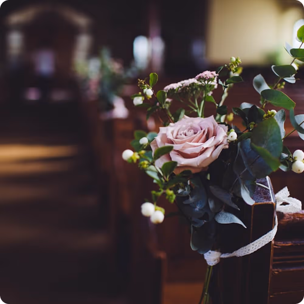 Close-up of a pink rose and greenery tied with a white ribbon to the end of a wooden church pew.