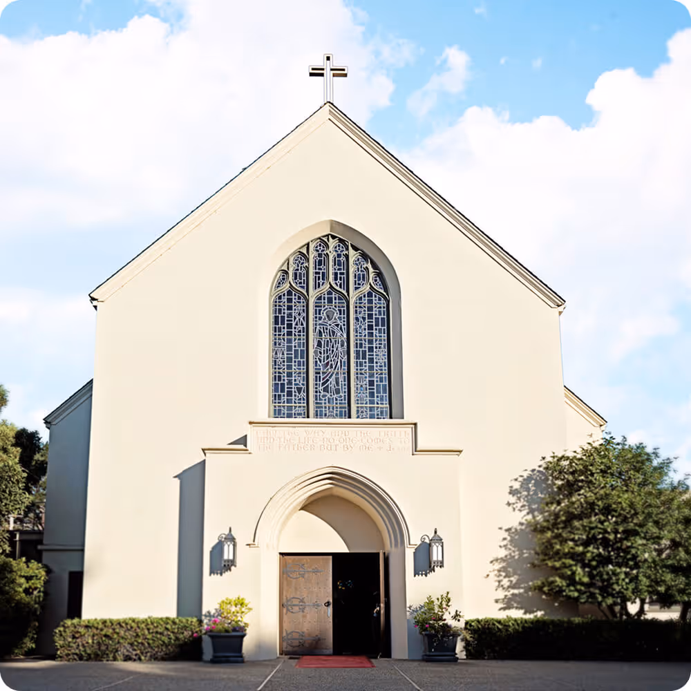 Front view of a beige church building with a wooden door, arched stained glass window, and a cross on the roof peak.