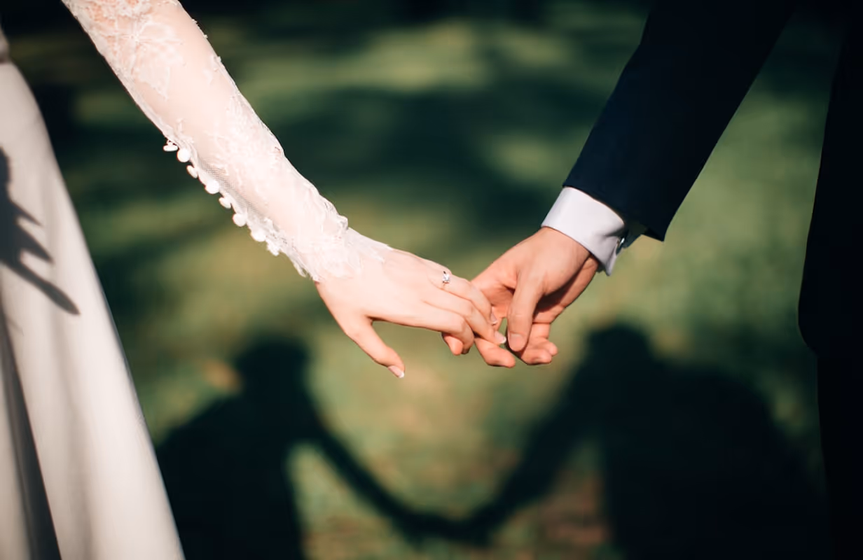Close-up of a bride and groom holding hands, with the bride wearing a laced wedding dress and an engagement ring.