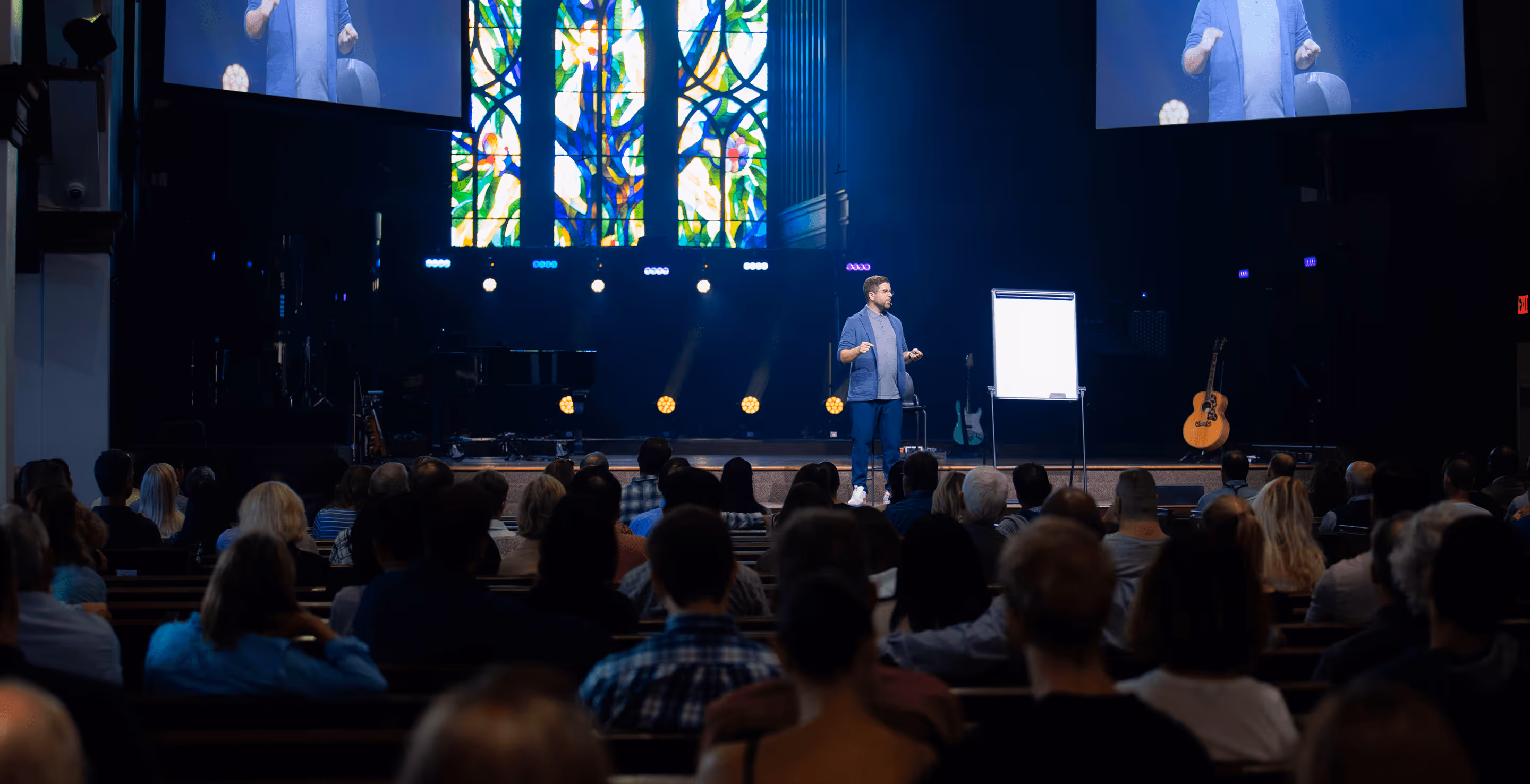 Speaker giving a presentation to an audience in a dark auditorium with stained glass windows and musical instruments on stage.