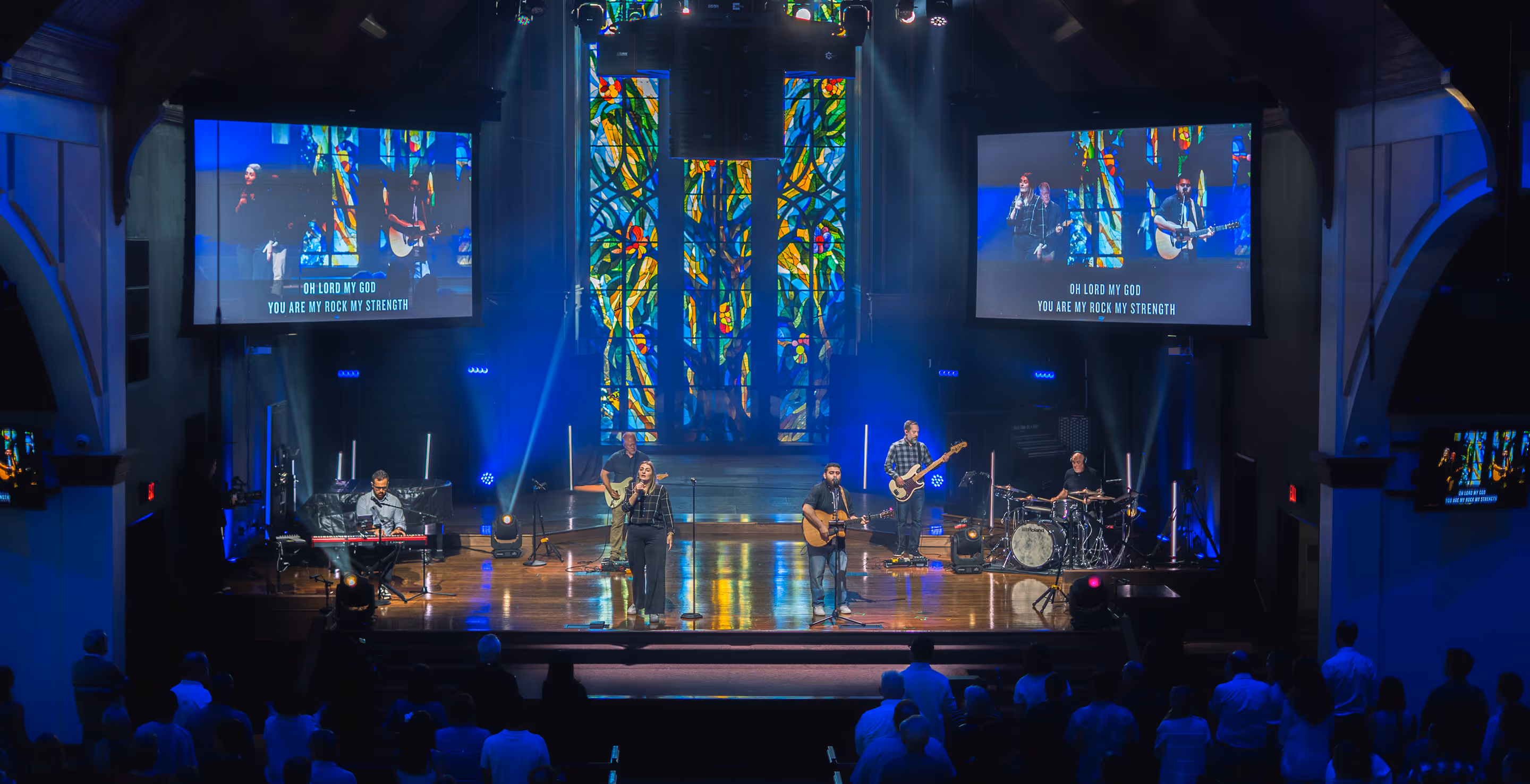 Worship band performs on stage with stained glass backdrop and lyrics shown on screens.
