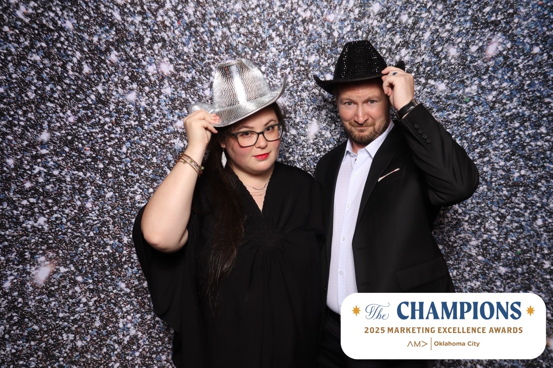 Two Hammonds Media & Marketing founders smiling in front of a sparkling backdrop at the 2025 AMA OKC Marketing Excellence Awards, wearing formal attire and playful event accessories.