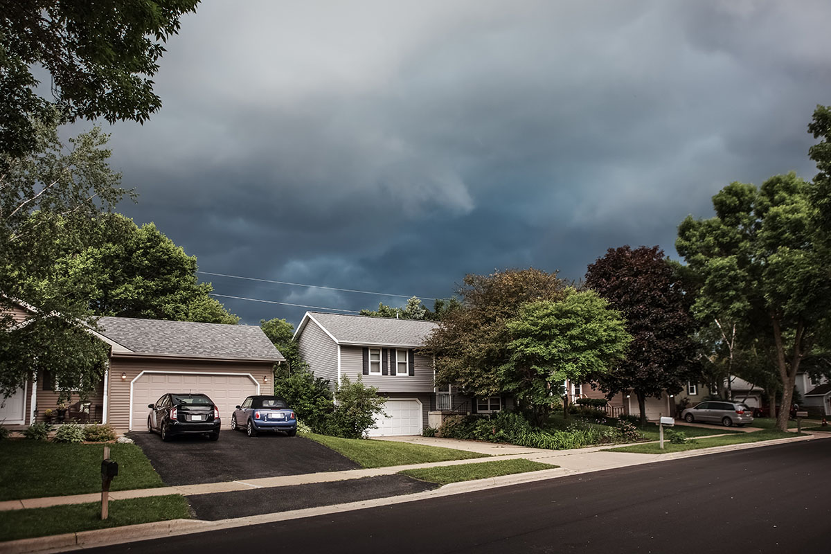 Dark storm clouds loom over a residential street with homes, cars, and trees, highlighting the need for roof preparation before summer storms.