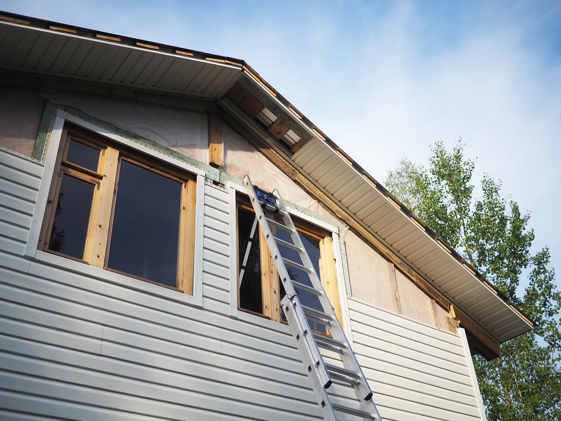 House exterior during siding replacement with partial vinyl siding removed, exposing house wrap, and ladder leaning against the gable.