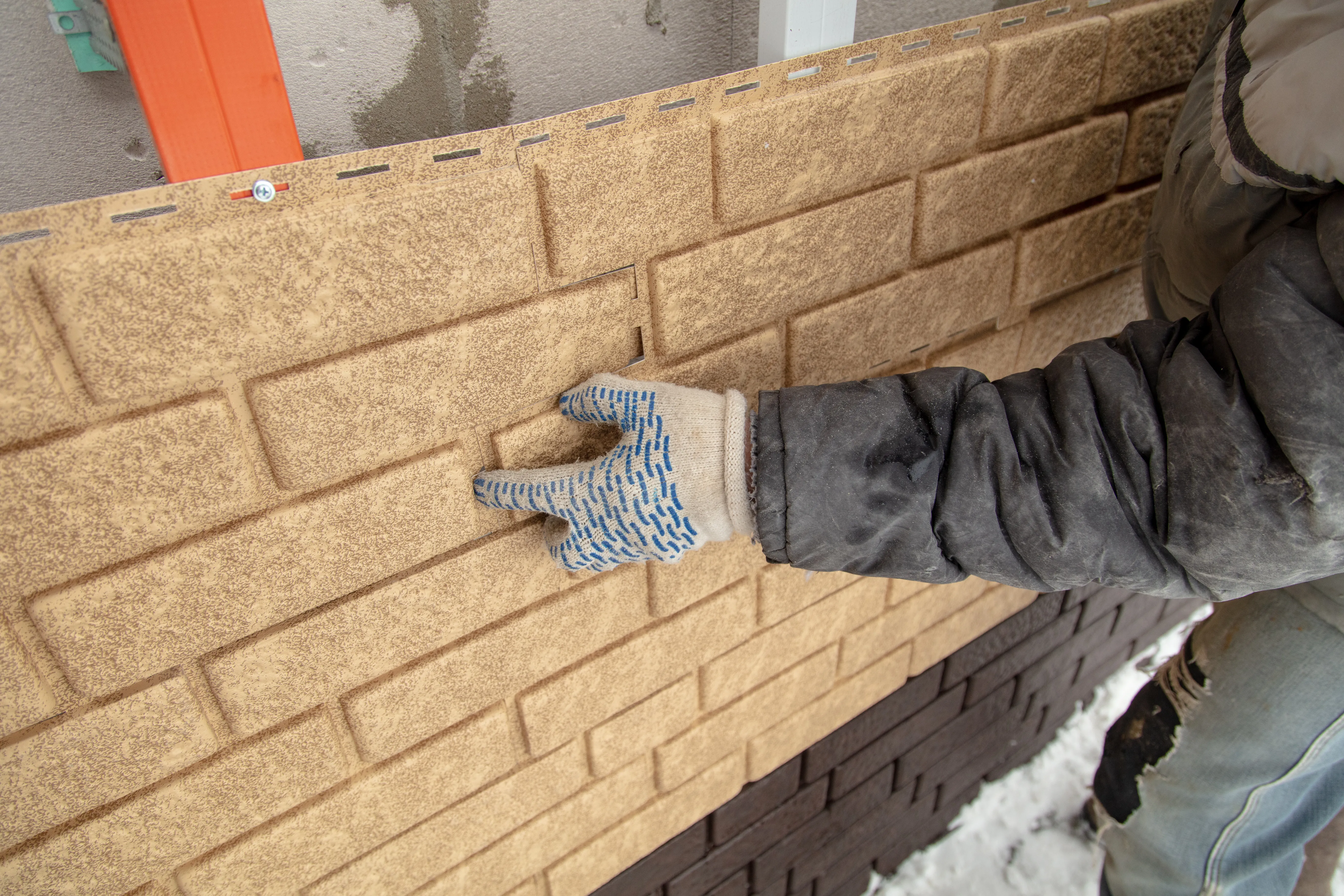 Worker in winter jacket and gloves touching newly installed faux brick vinyl siding on house exterior.