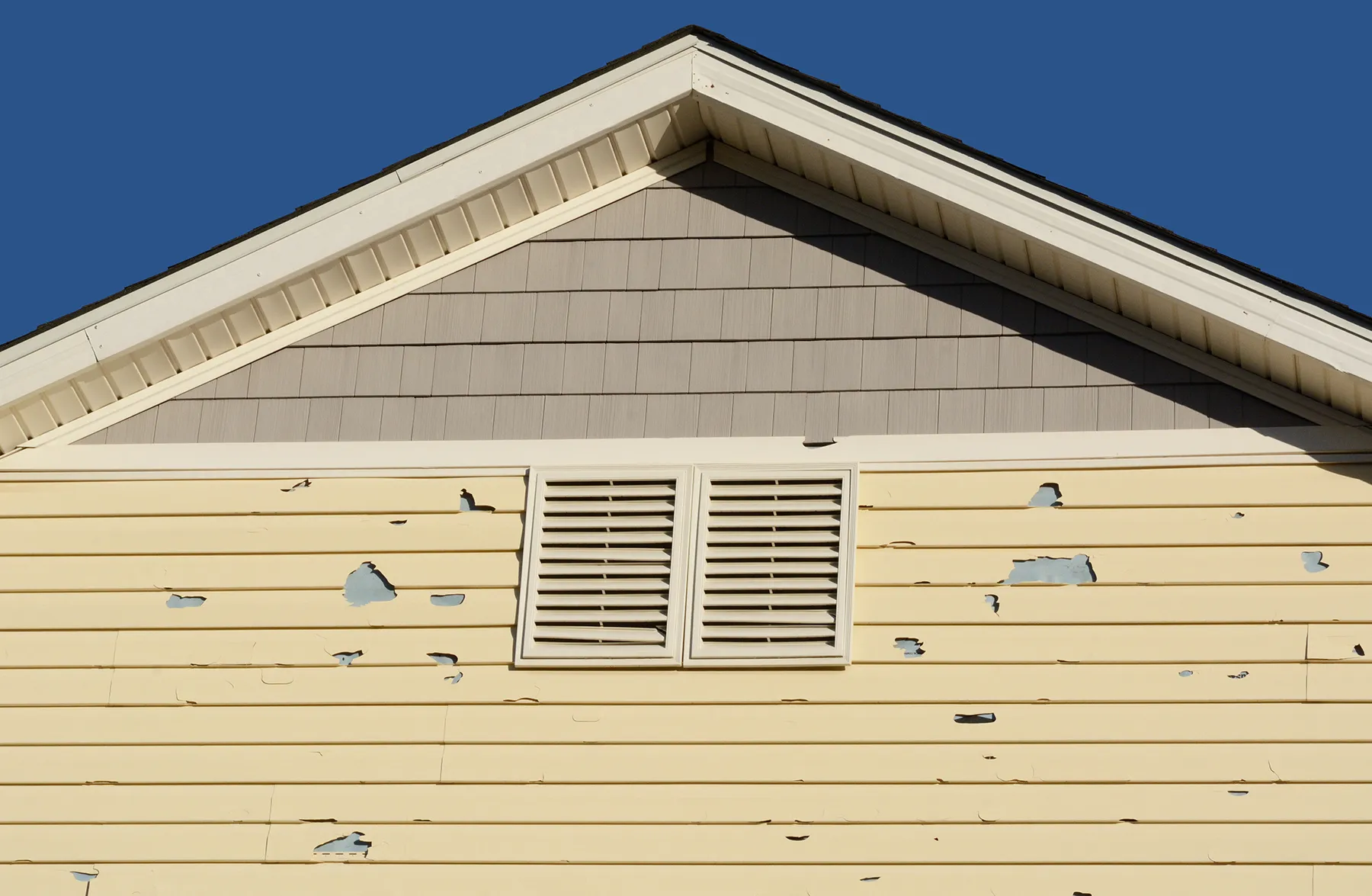 Close-up of house gable with peeling and chipped yellow vinyl siding around louvered vents, showing signs of wear.