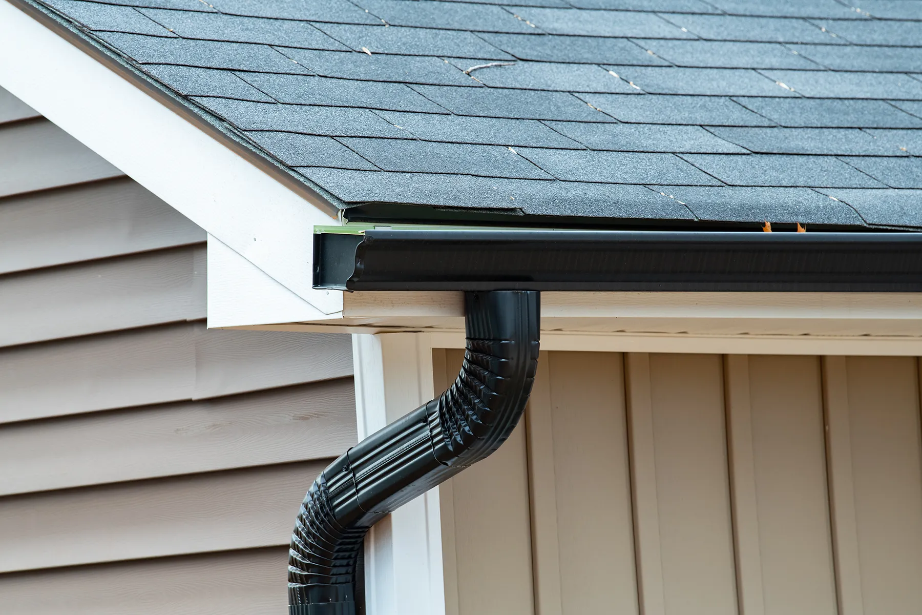 Black aluminum gutter and corrugated downspout installed on house with gray shingles and vinyl siding.