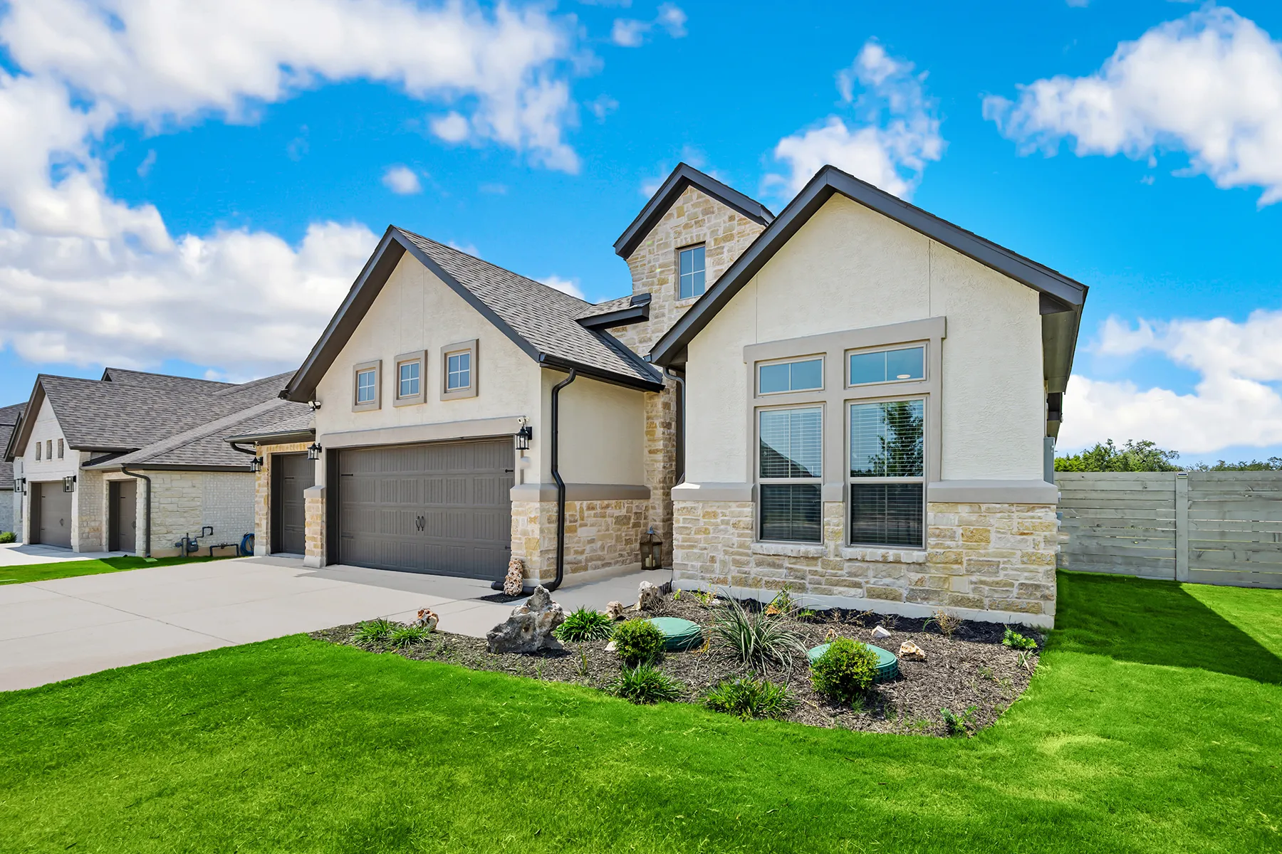 Modern suburban home with beige stucco and stone exterior, attached garage, and landscaped yard under blue sky.