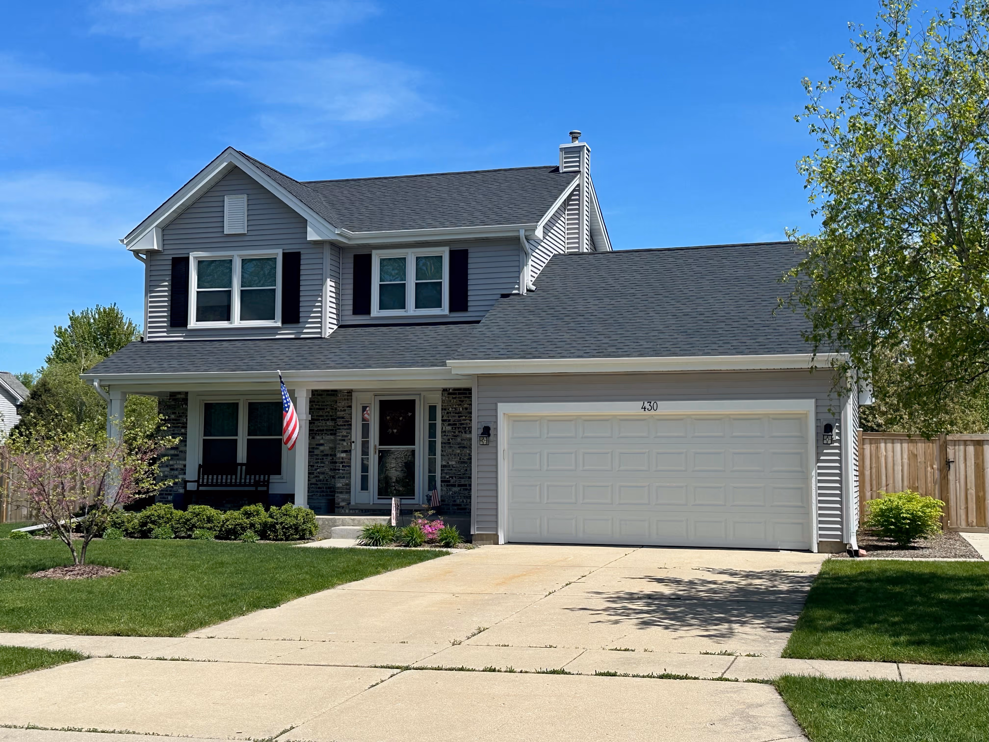 Two-story gray vinyl-sided home with black shutters, stone-accented porch, attached garage, and dark asphalt shingle roof on sunny day.