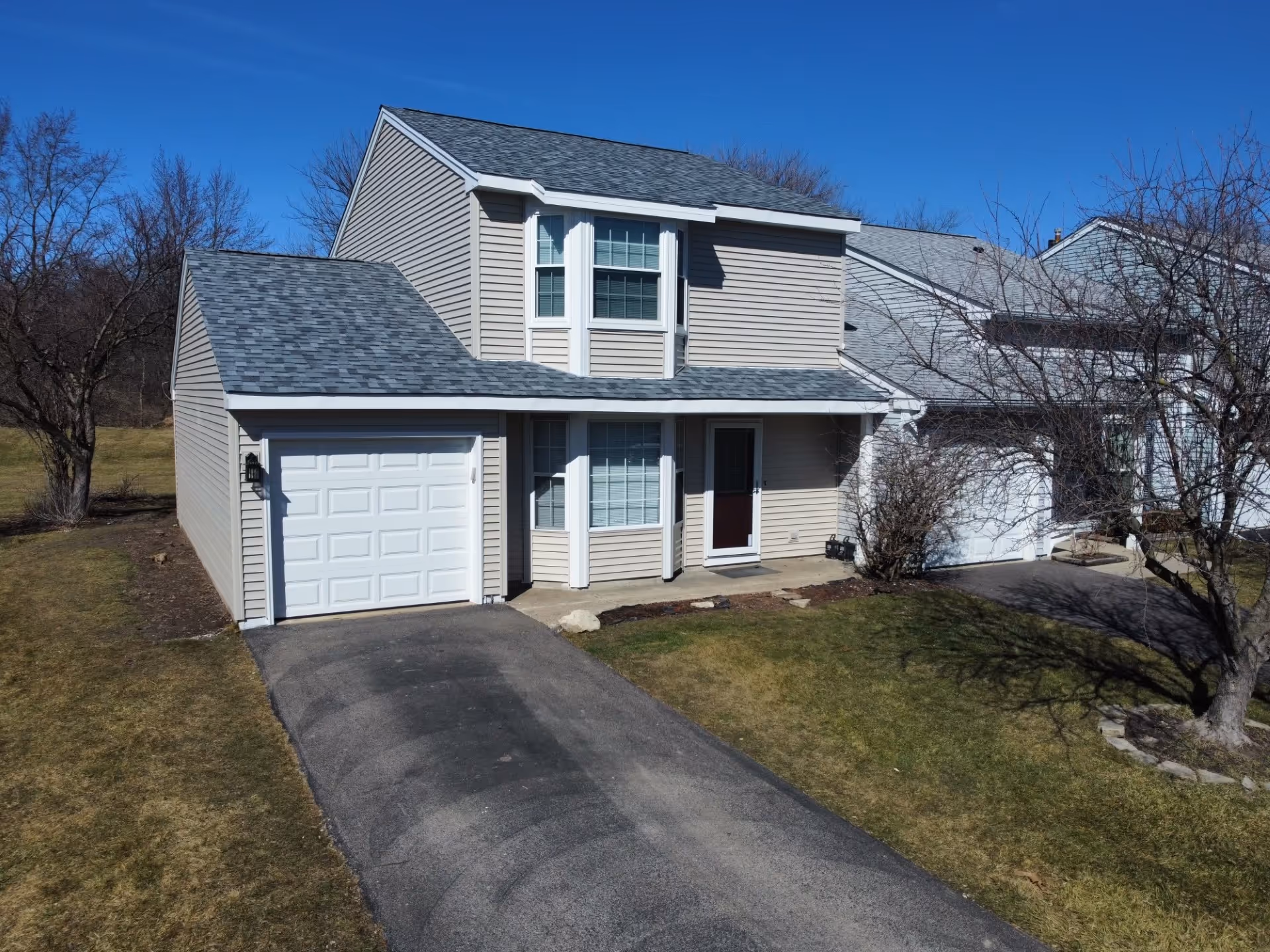 Two-story beige vinyl-sided home with attached garage, bay window, covered entry, and gray asphalt shingle roof on sunny day.