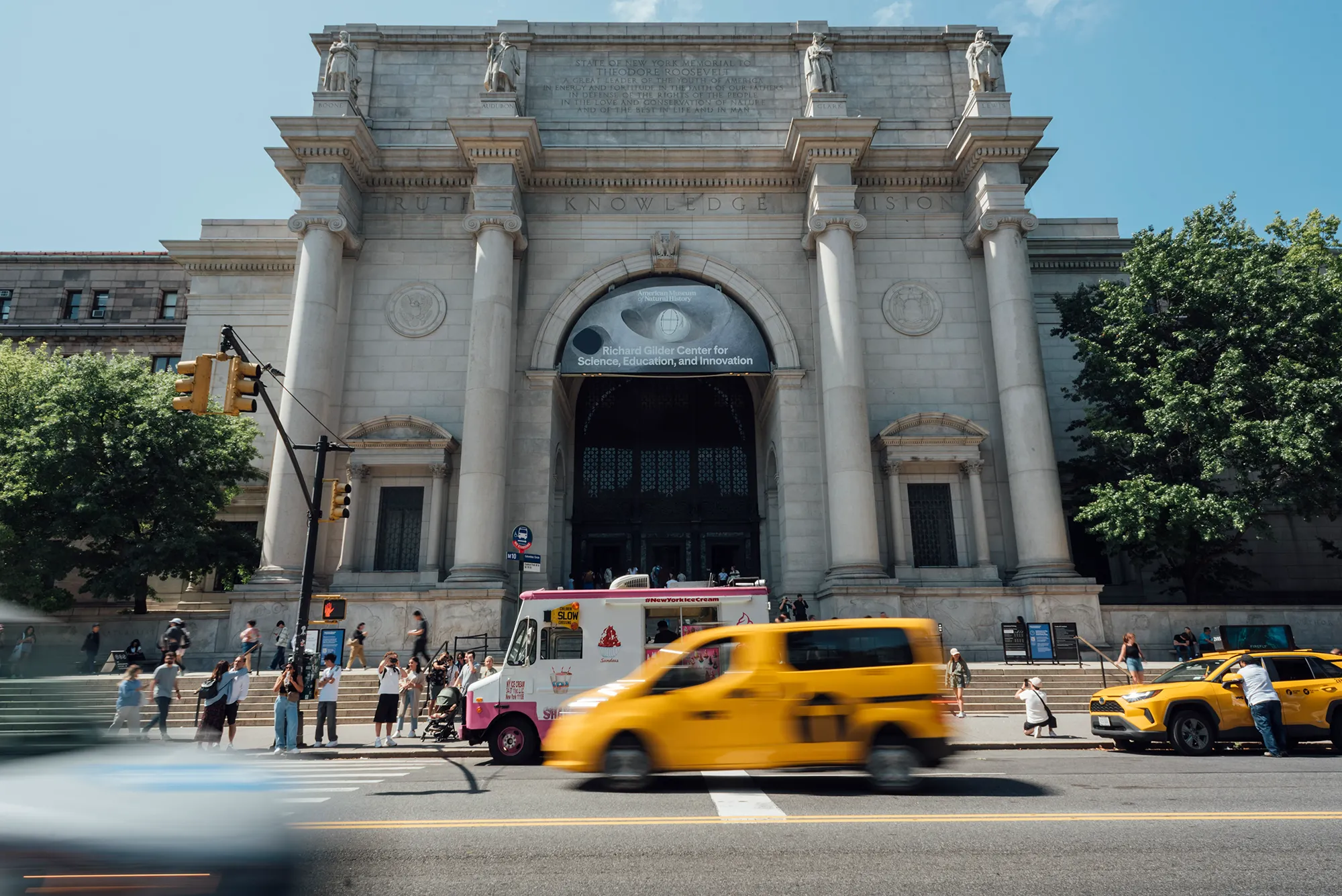 A yellow taxi drives along a city street, surrounded by buildings and other vehicles.