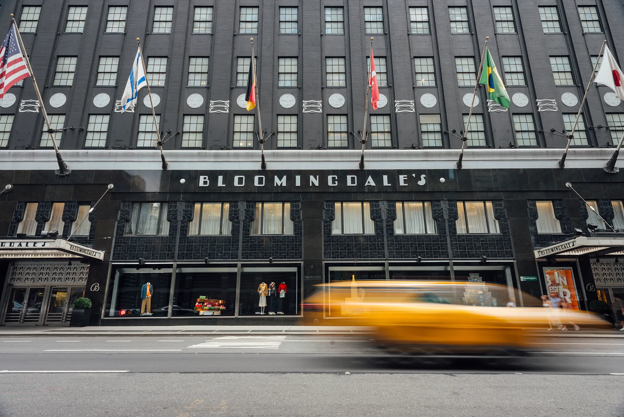 A yellow taxi drives past a building adorned with multiple flags, showcasing a vibrant urban scene.