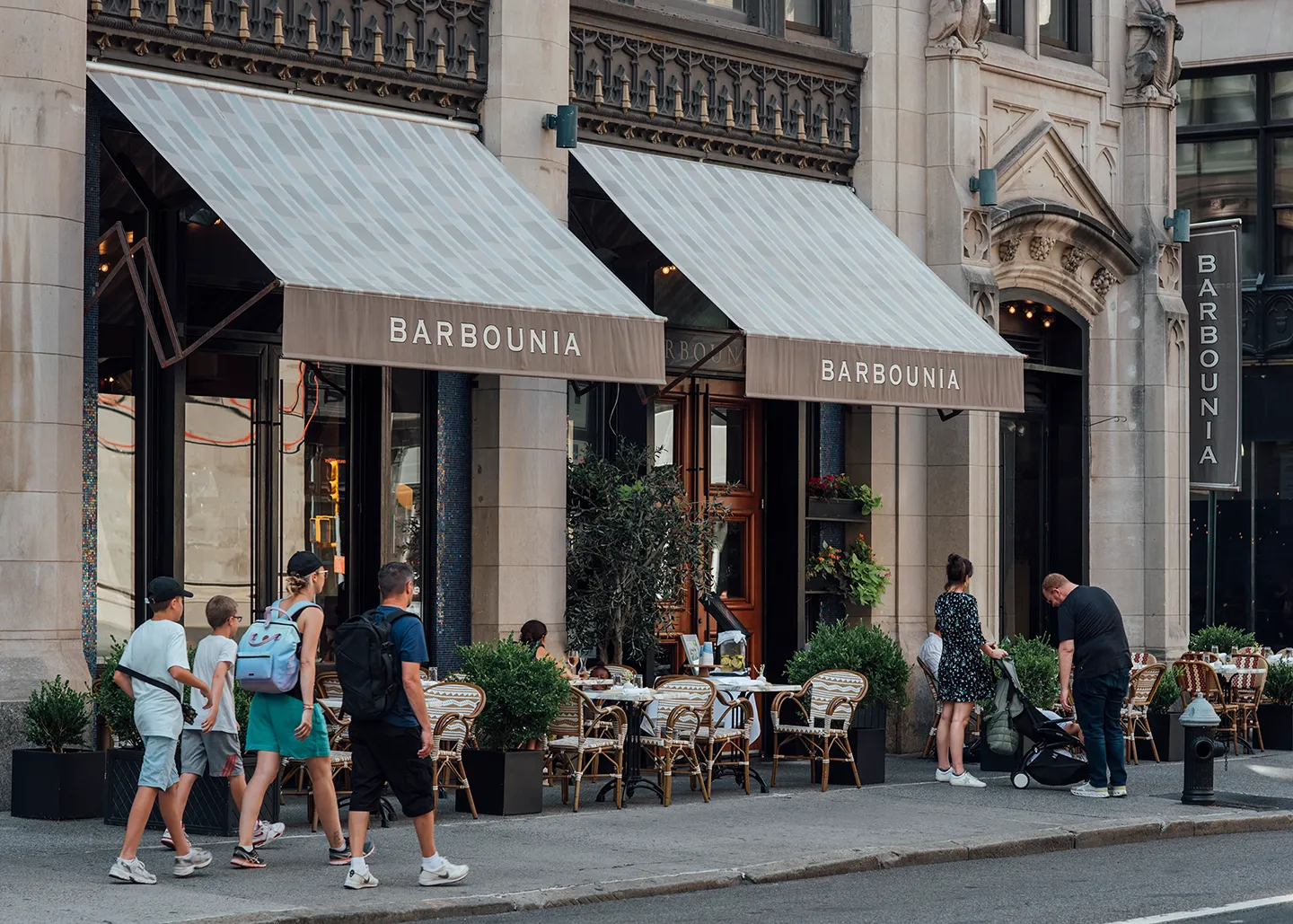 A group of people walking on the sidewalk in front of a restaurant, engaged in conversation and enjoying the day.