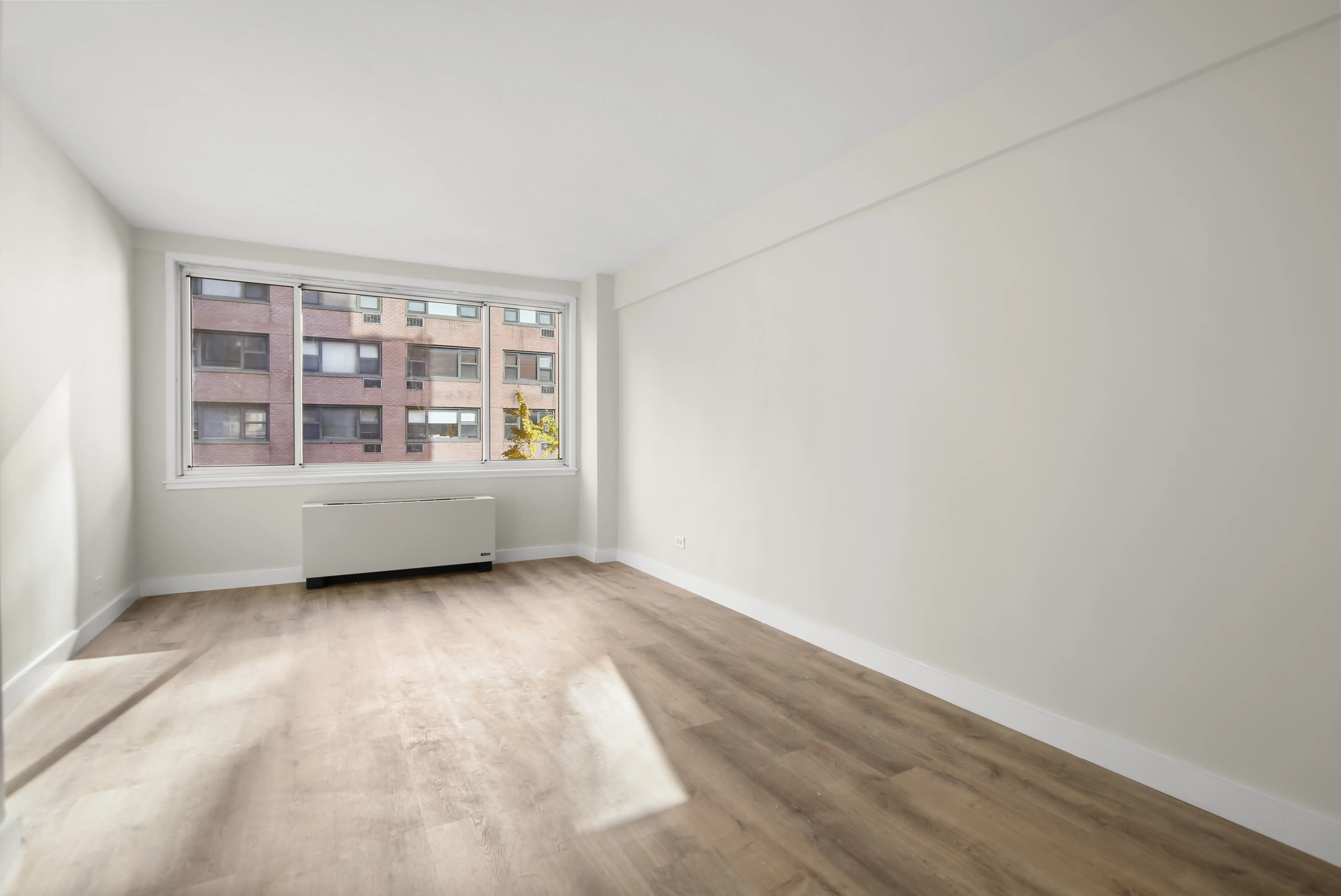 A vacant room with polished hardwood floors and a window, creating a bright and airy atmosphere.