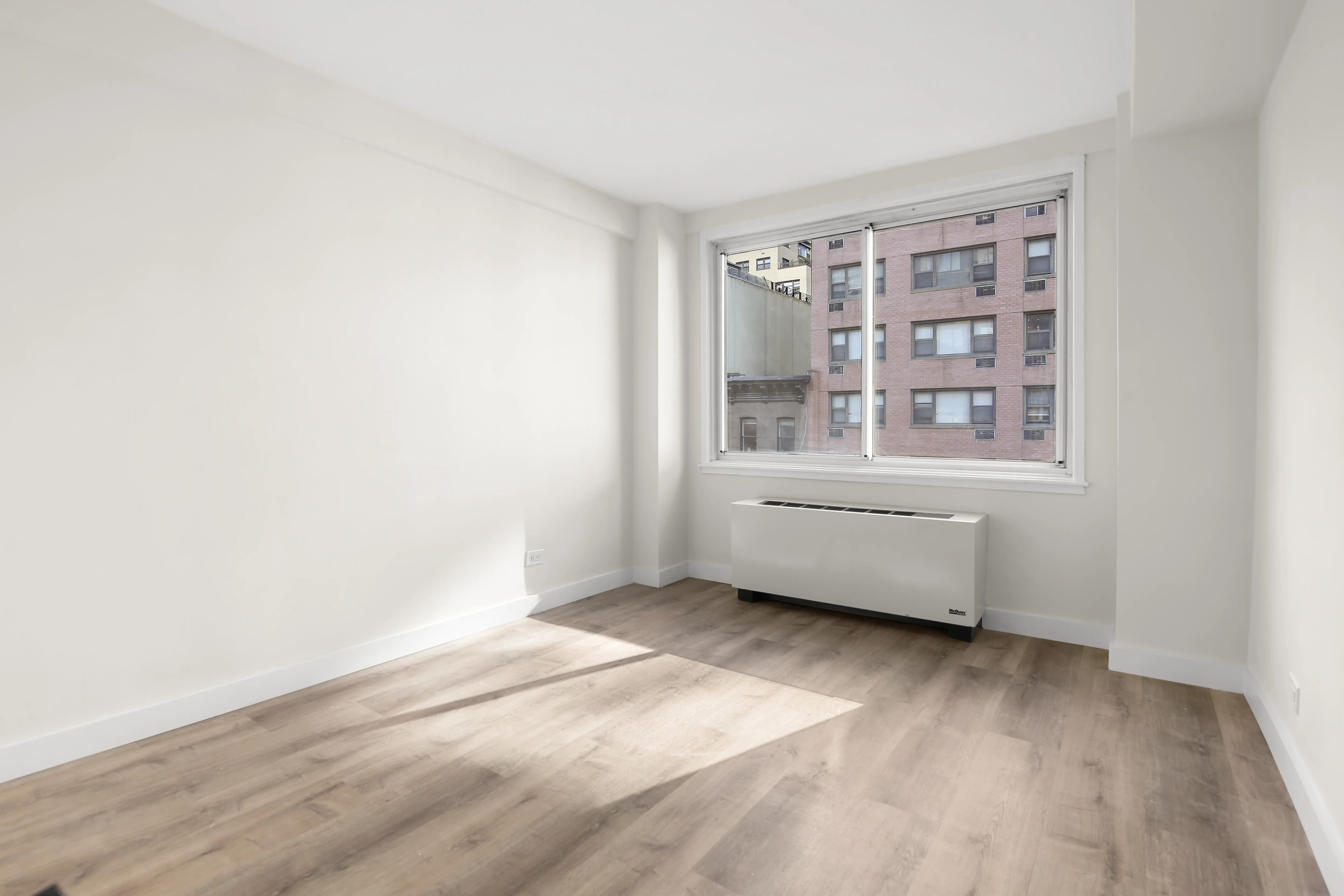 A vacant room with polished hardwood floors and a window, creating a bright and airy atmosphere.