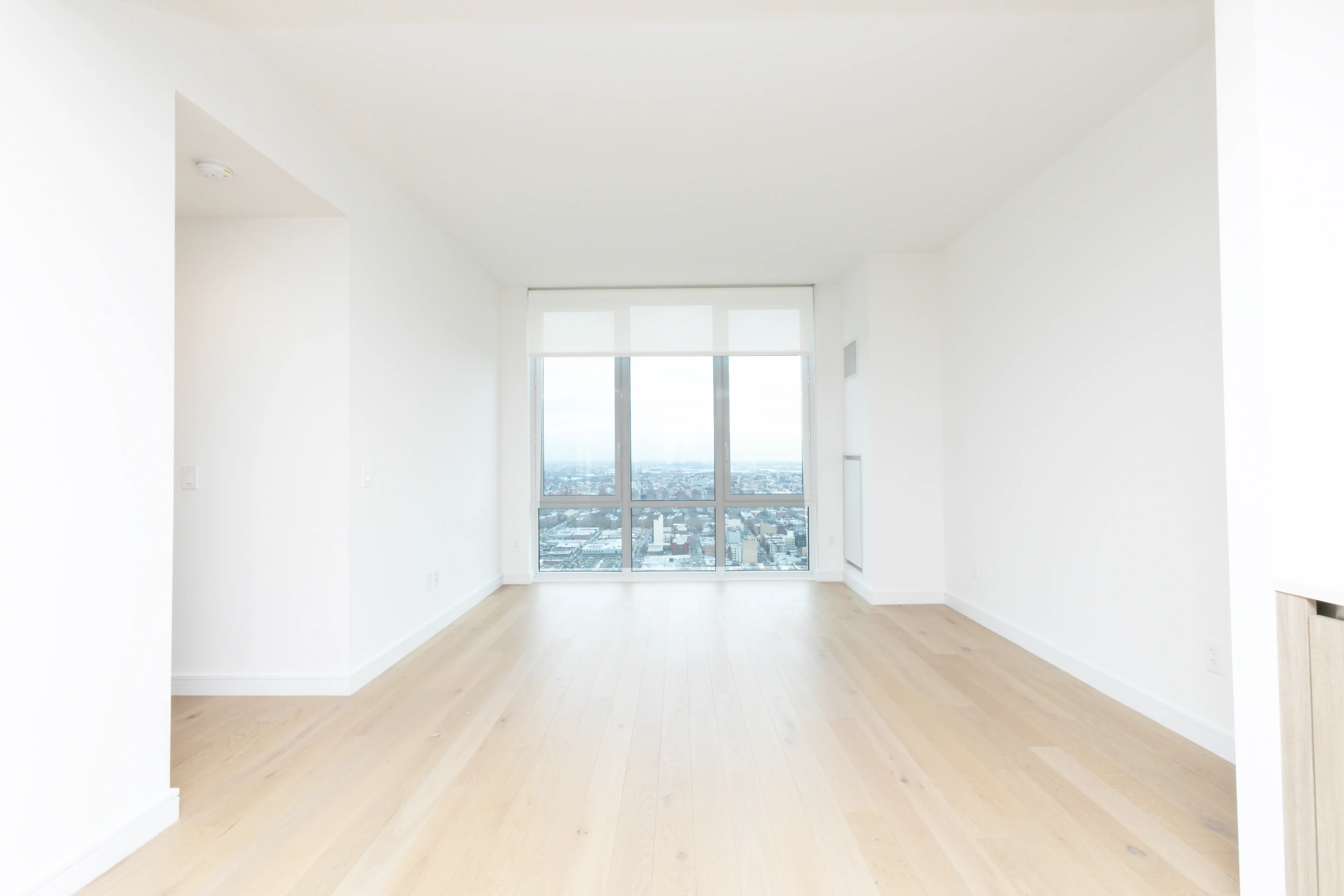 A bright white room with hardwood floors, featuring a large window that offers a view of the city skyline.
