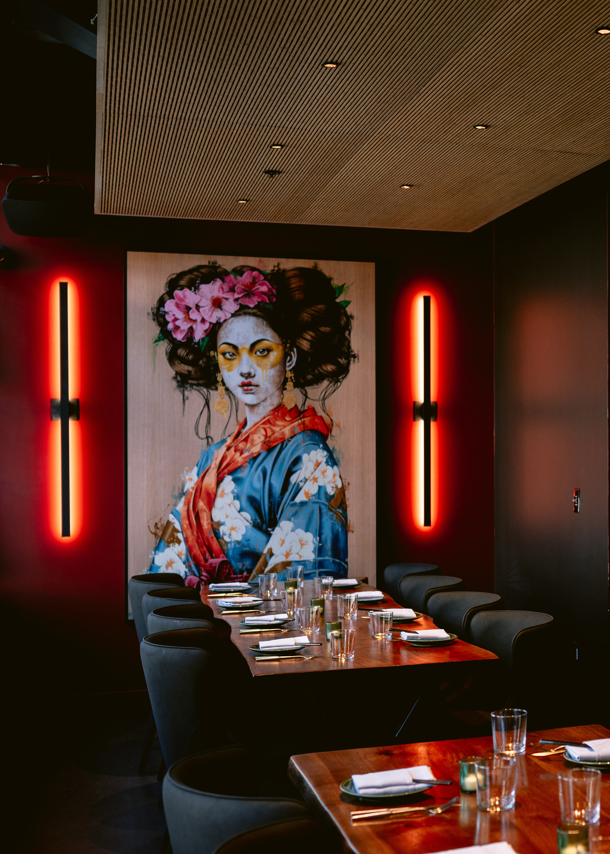 Interior view of Kaiyō restaurant featuring a long dining table with grey chairs, under warm pendant lighting. The room has a vibrant red wall accented by decorative wooden panels with circular motifs