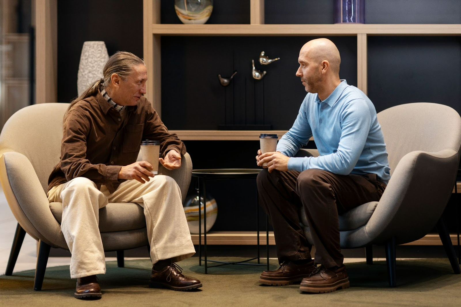 Two men sitting in modern chairs at a wellness clinic, having a supportive conversation over coffee, symbolizing community and care during TRT therapy.