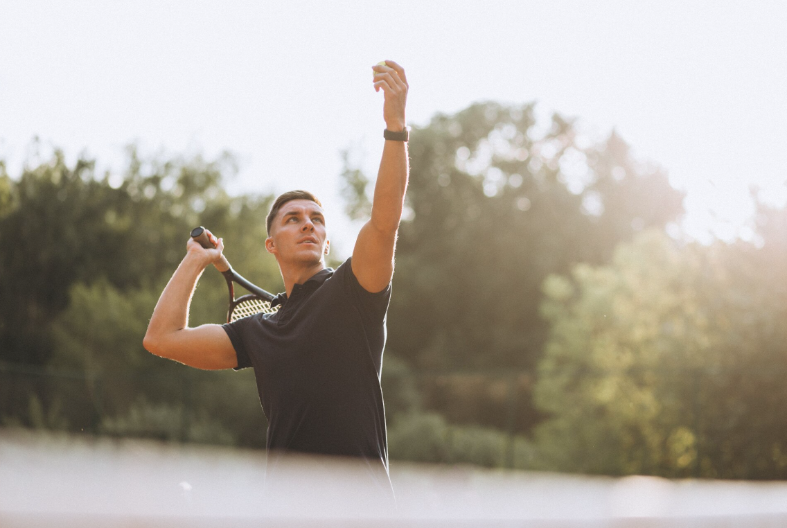 A man prepares to serve a tennis ball outdoors, representing increased energy, strength, and focus as signs of progress from TRT treatment