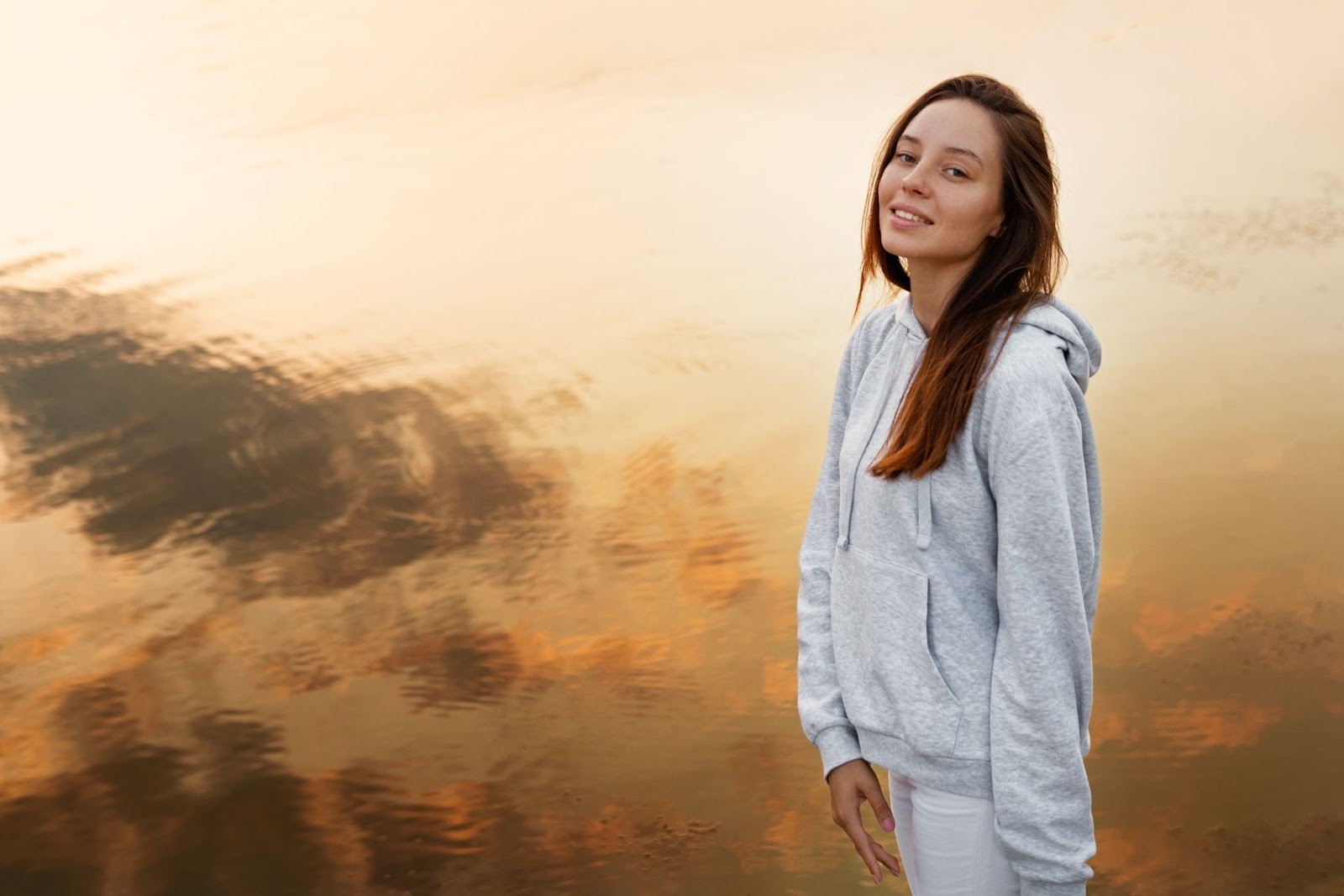 A woman wearing a hoodie stands beside a tranquil body of water, reflecting on the peaceful surroundings