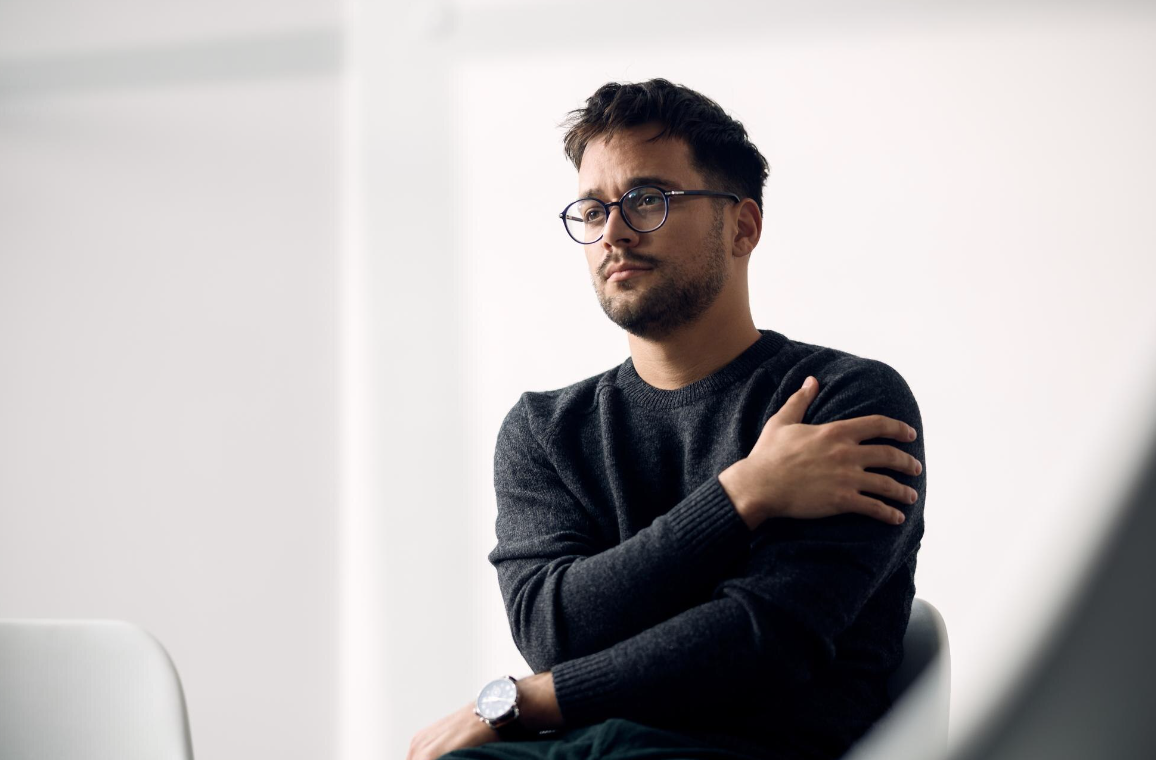 A man with glasses sits comfortably on a chair, appearing relaxed and contemplative