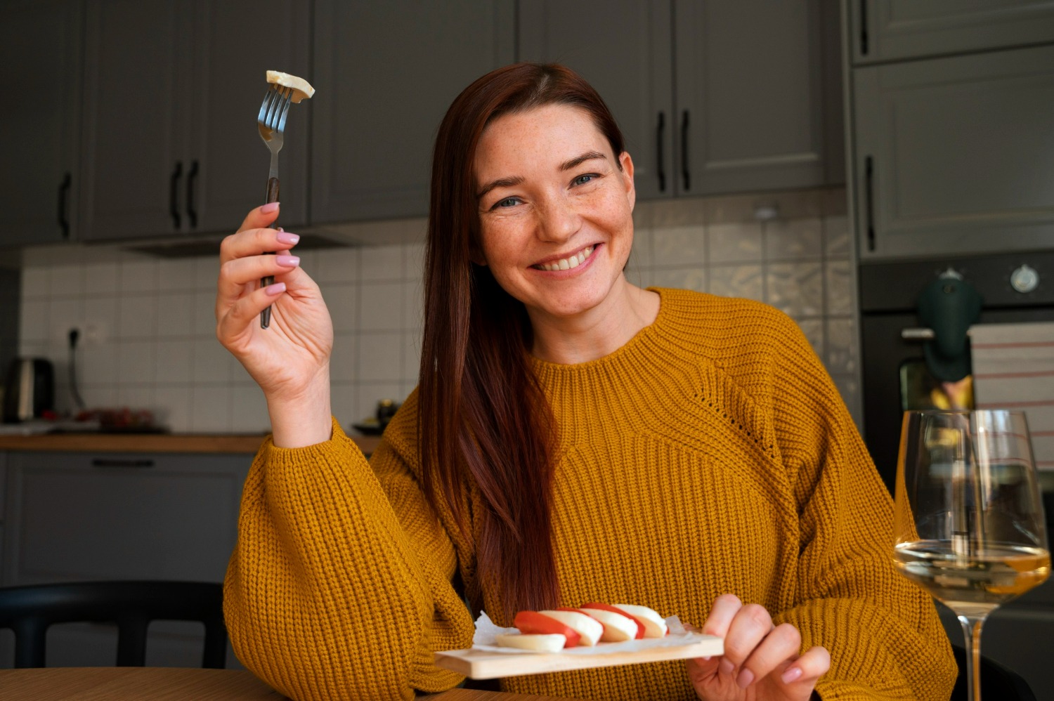 A smiling woman wearing a mustard top, looking at the camera while eating a healthy meal in a kitchen
