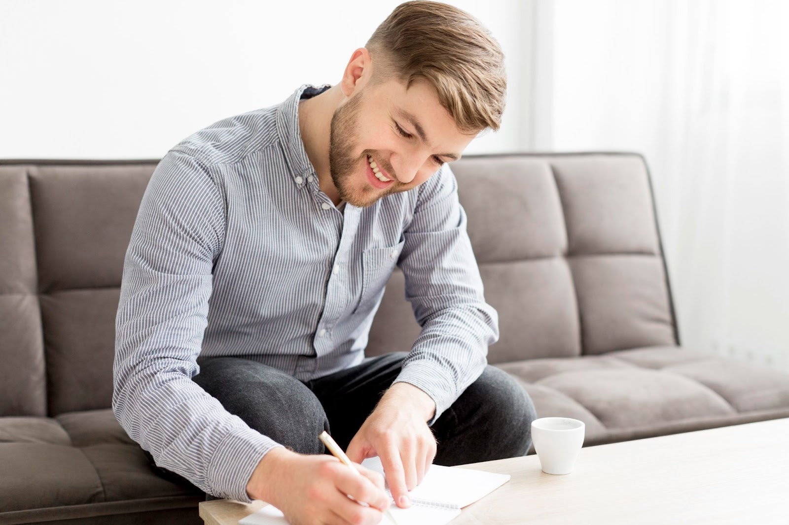 A man smiling, sitting on a couch, and writing something down on a notebook