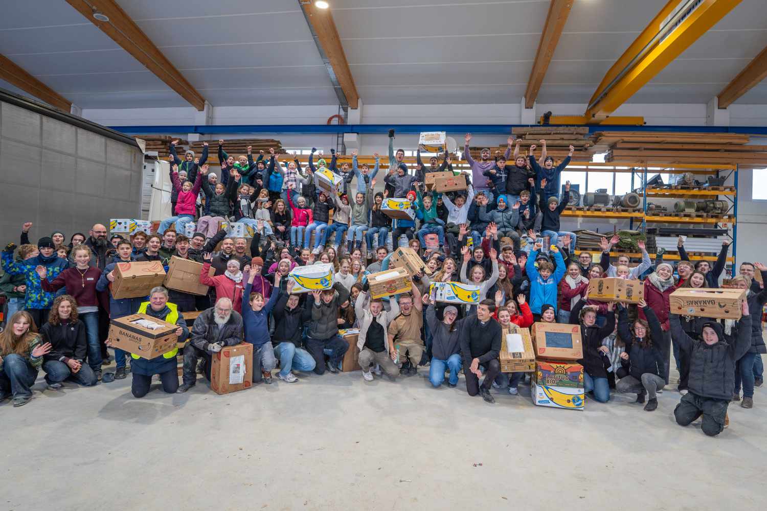 Große Gruppe von JLH-Helfern in einer Lagerhalle mit gesammelten Hilfspaketen