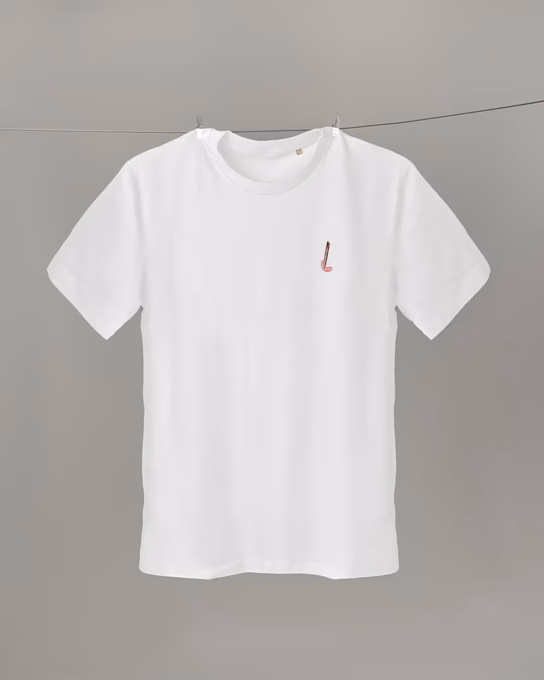 Ein weißes T-Shirt hängt an einer Leine vor grauem Hintergrund. Auf der linken Brust ist eine kleine Stickerei einer grünen Pflanze mit braunem Stängel angebracht, die für ein schlichtes, natürliches Gefühl sorgt.