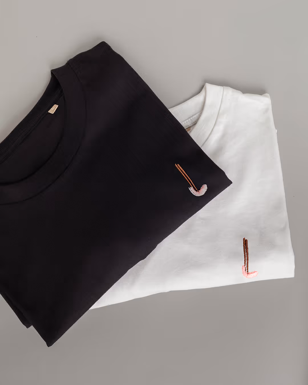 Zwei gefaltete T-Shirts auf grauem Untergrund: ein schwarzes, ein weißes, jeweils mit einer kleinen rosa Musiknote bestickt. Die Szene wirkt minimalistisch und stilvoll.