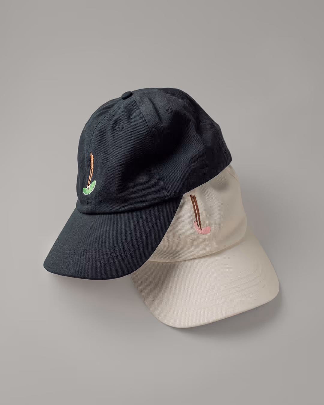 Zwei Kappen überlappen sich auf neutralem Hintergrund, eine in Schwarz und eine in Beige. Jede ist mit einem kleinen gestickten Golfschläger-Design versehen, das ihr einen sportlichen und minimalistischen Touch verleiht.