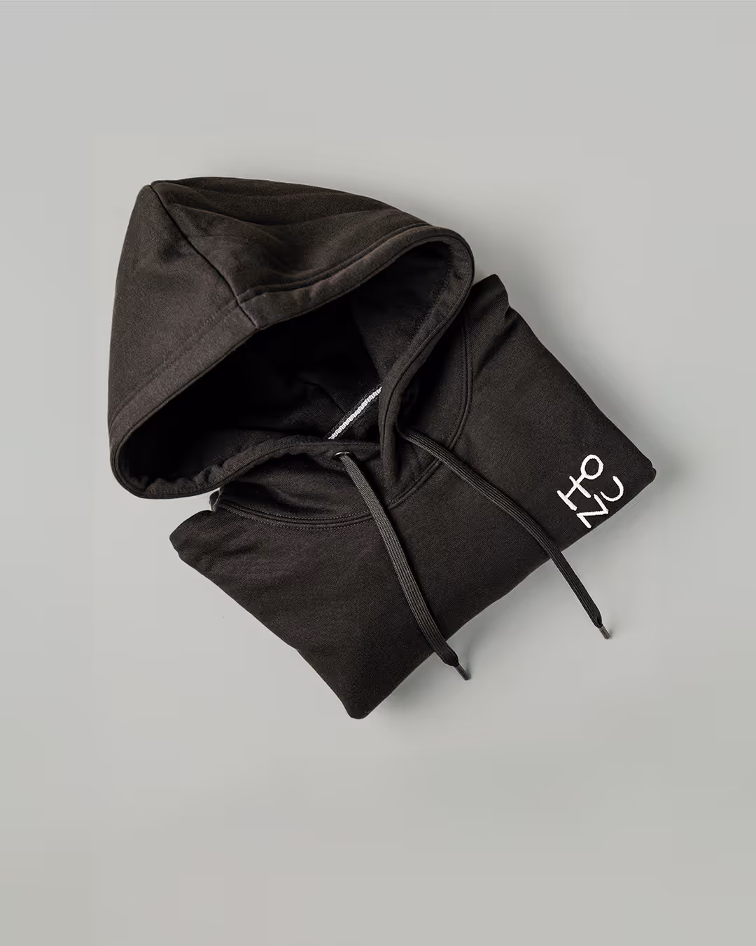 Ein ordentlich gefalteter schwarzer Hoodie mit Kapuze und Kordelzug, mit weißem „TONU“-Schriftzug auf dem rechten Ärmel. Auf hellgrauem Hintergrund.