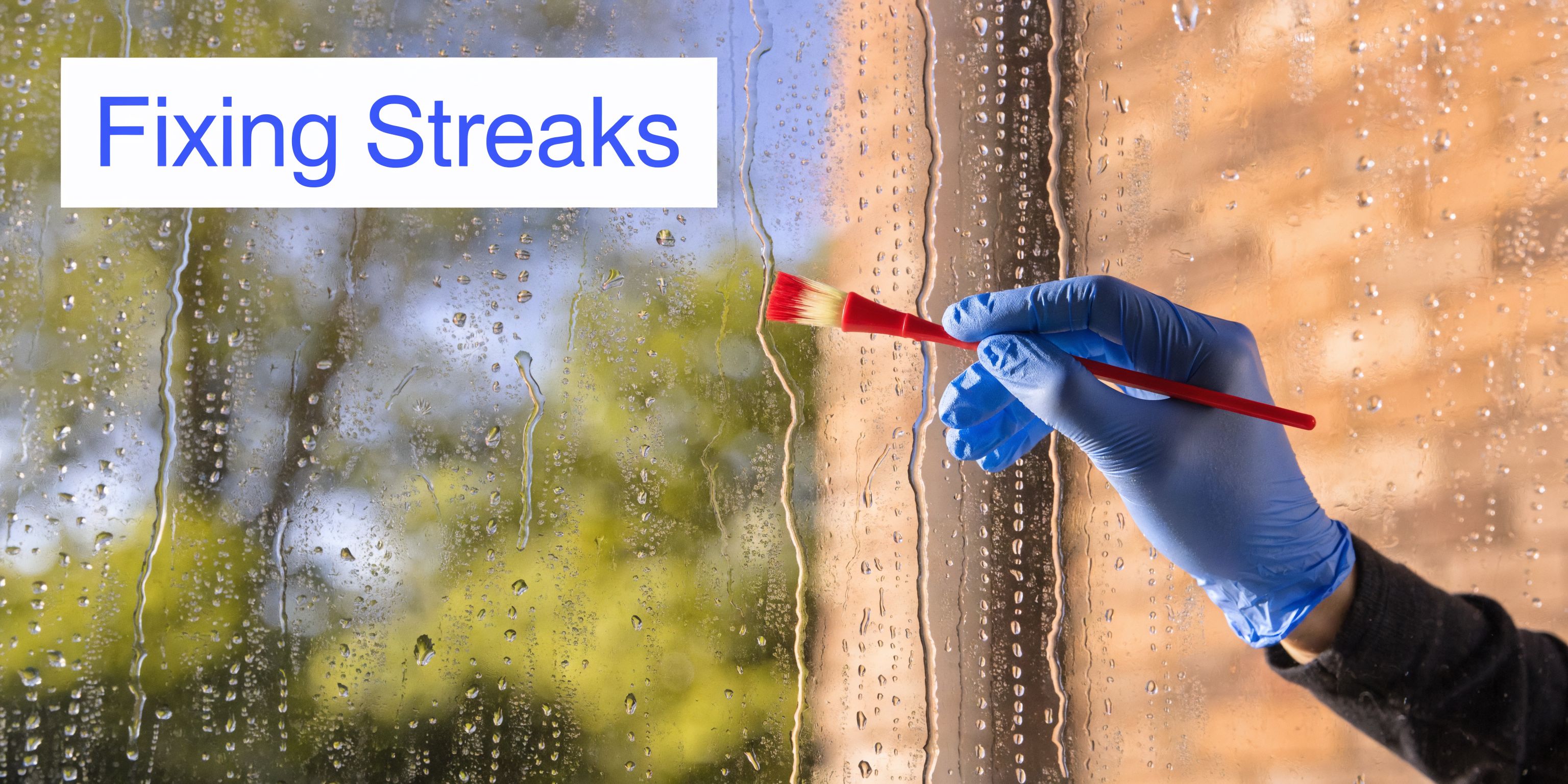 A person wearing a blue glove uses a small red brush to clean streaks on a wet glass window.