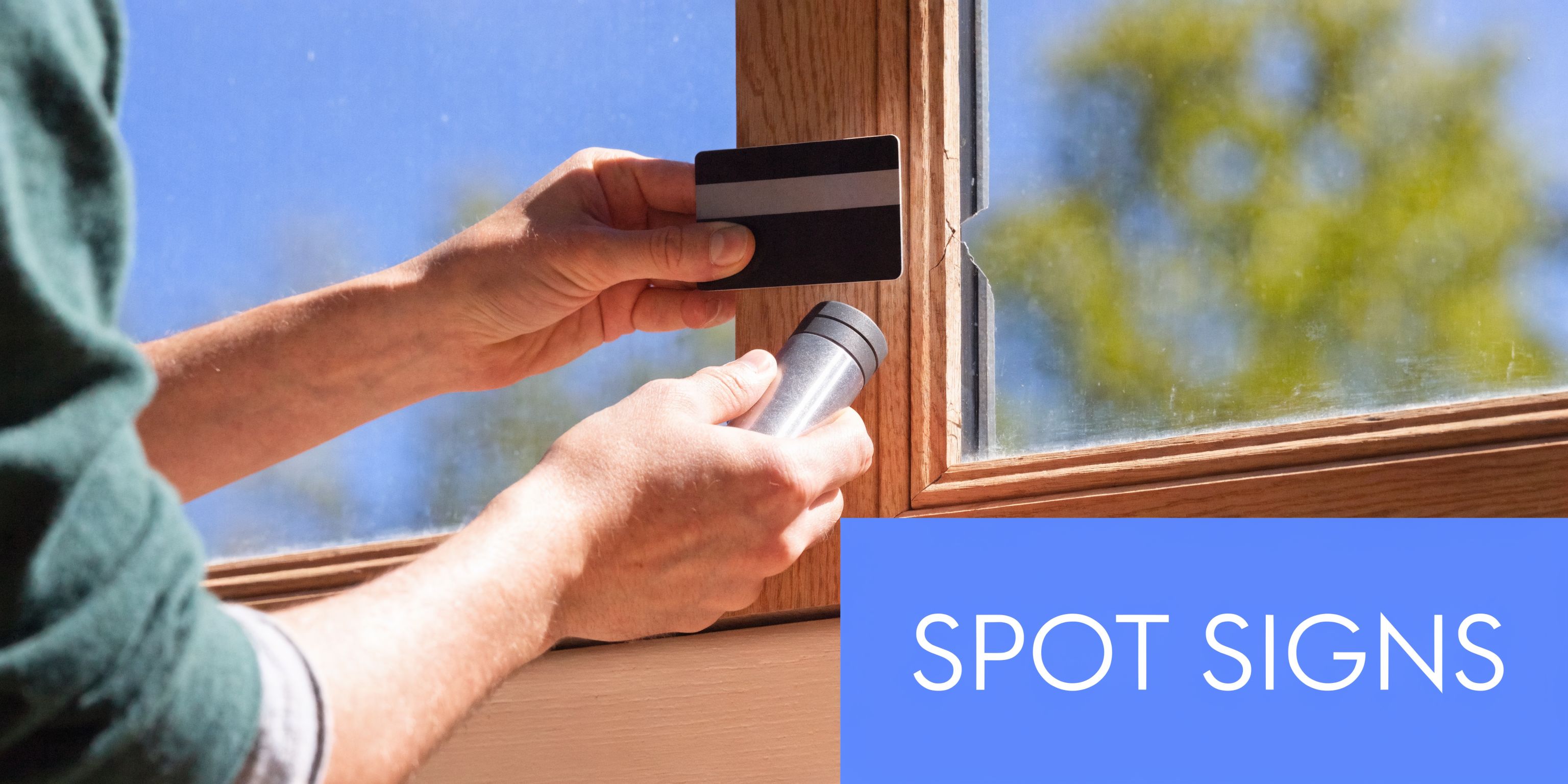 A person holding a credit card and a small flashlight to inspect a wooden window frame seal.