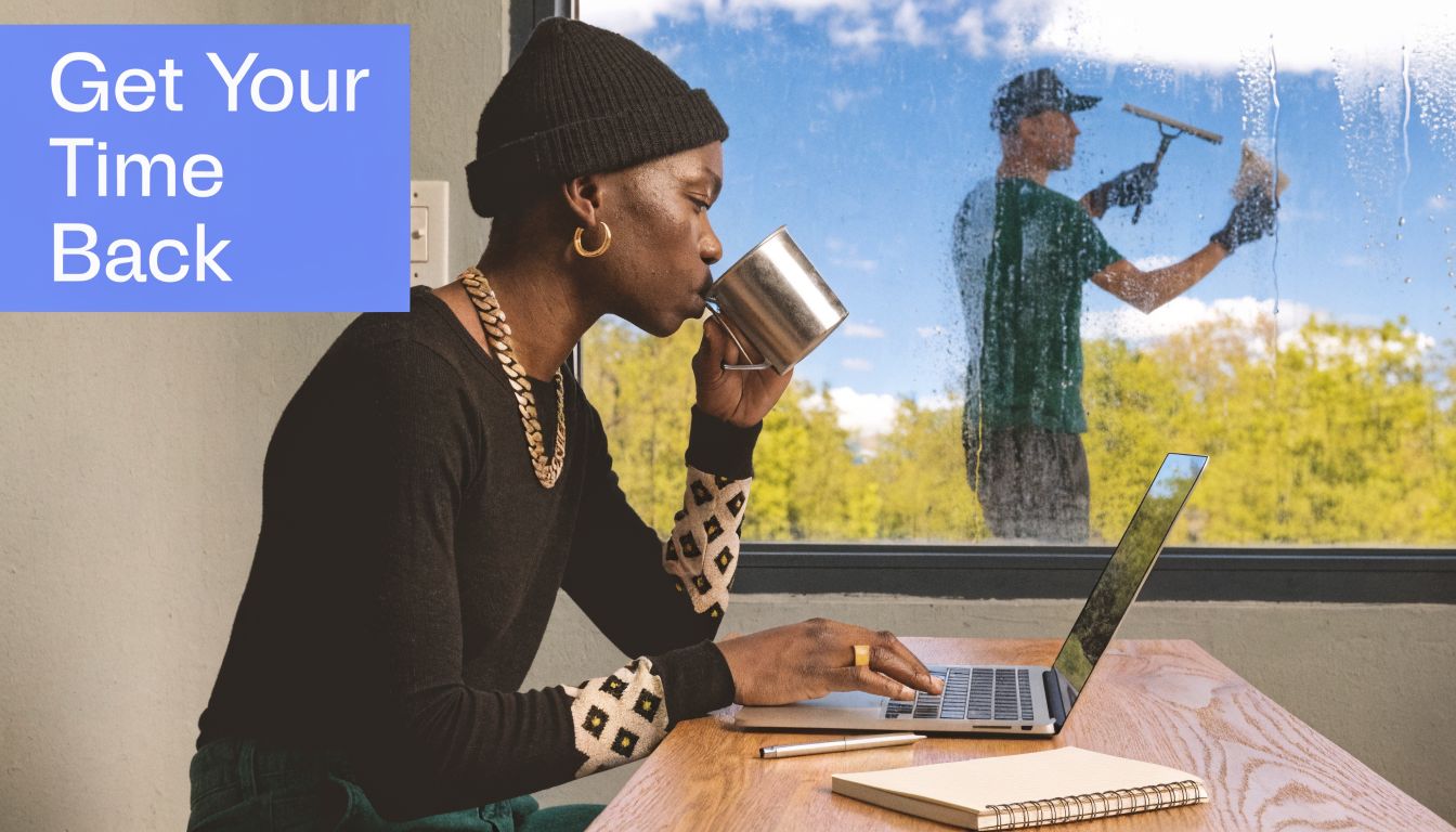 A person working on a laptop while drinking coffee as a professional cleaner washes the window.