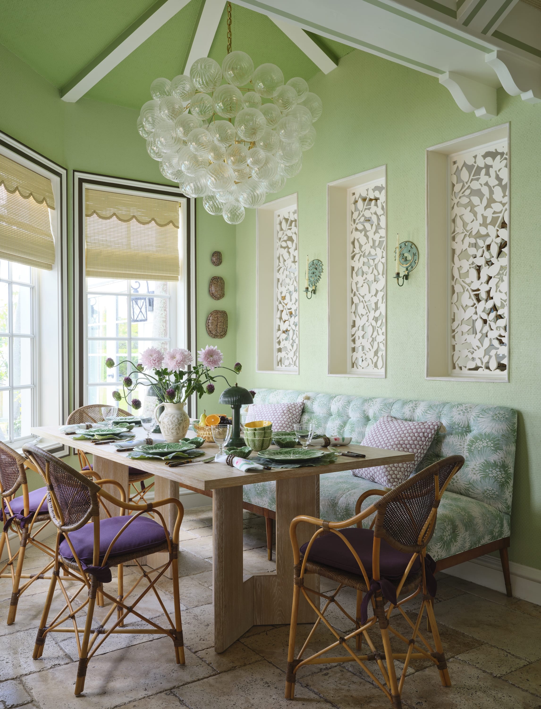 Cozy dining area with a wooden table set with green leaf-patterned plates, wicker chairs with purple cushions, a tufted bench with botanical upholstery, green walls, and a large glass bubble chandelier.