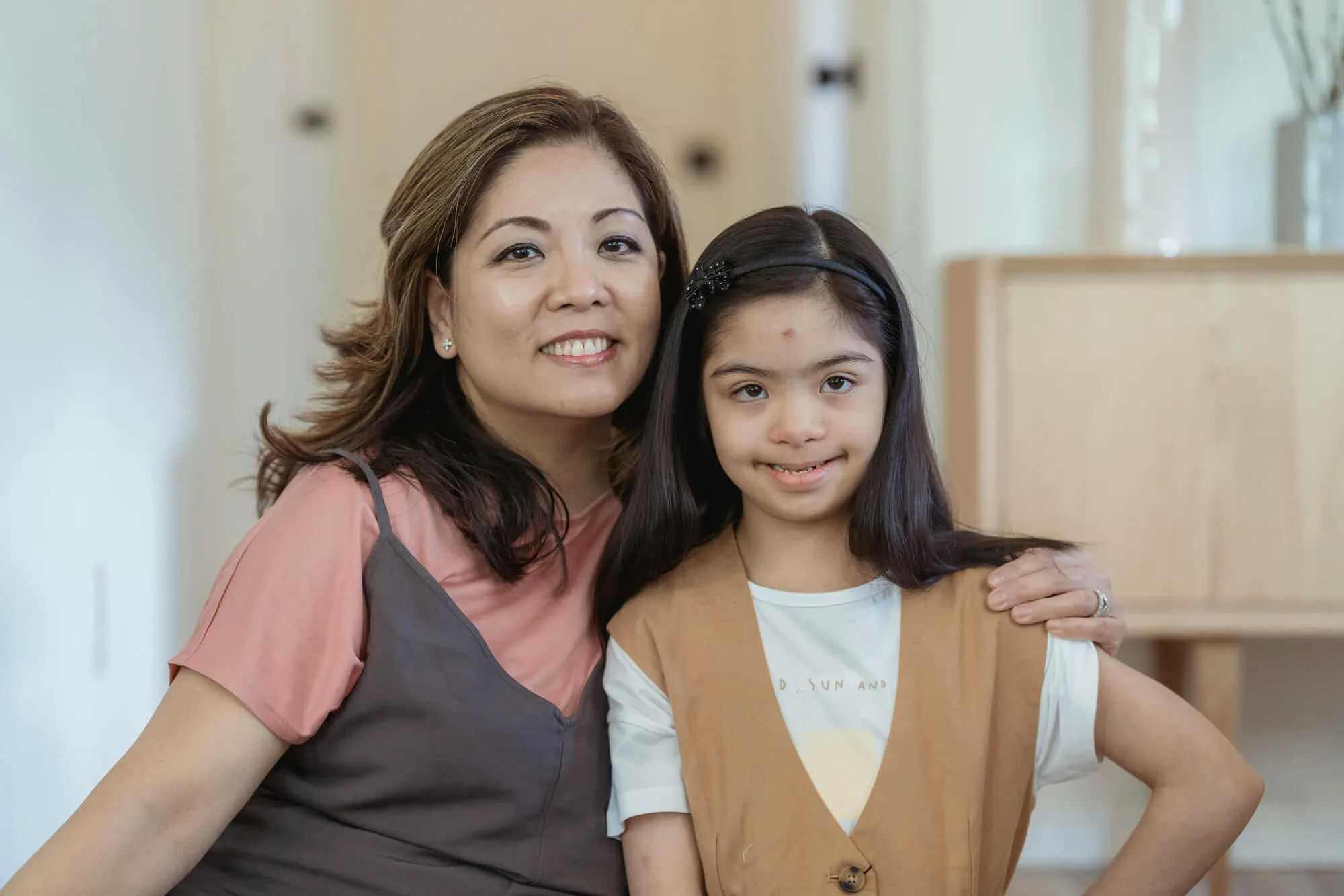 Mother and daughter smiling together at home, with the child displaying features associated with Down syndrome. The mother has her arm around her daughter, showing love, support, and inclusive family care. The image highlights themes of disability advocacy, family bonding, and pediatric healthcare awareness.