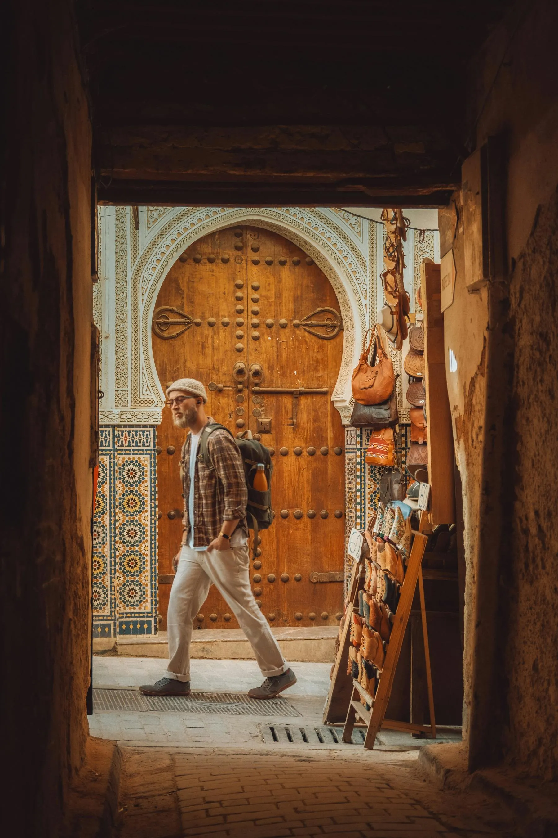 Traveler walking through a narrow alley in front of an ornate wooden Moroccan door, surrounded by traditional mosaic tiles and a display of handcrafted leather bags. The scene captures authentic Moroccan architecture and artisan culture in a historic medina.