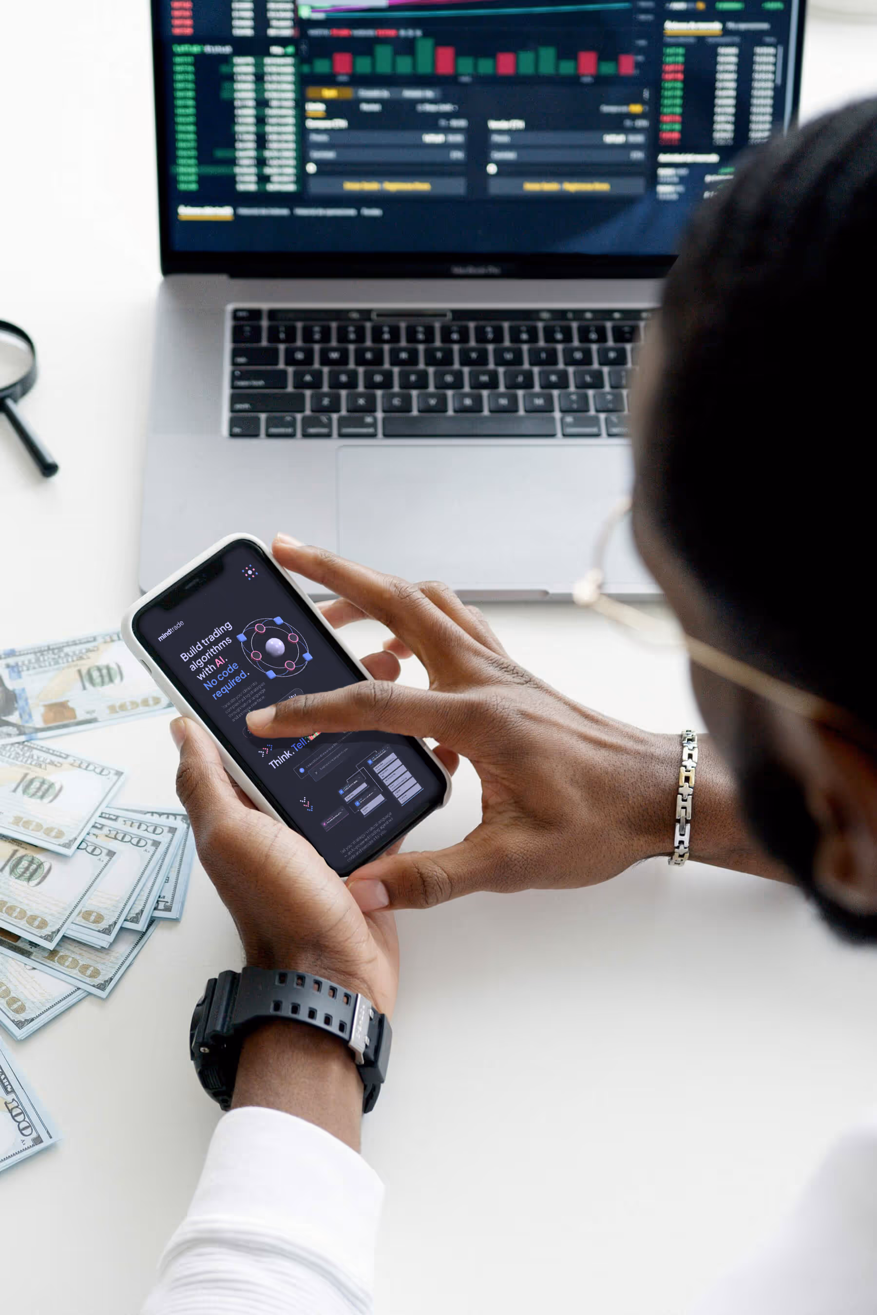 Trader using a crypto trading app on a smartphone with the MindTrade interface displayed, showing a no-code strategy builder. In the background, a MacBook displays live cryptocurrency charts and market data. U.S. dollar bills are stacked on the desk, emphasizing active investing, algorithmic trading, and mobile-first financial technology.